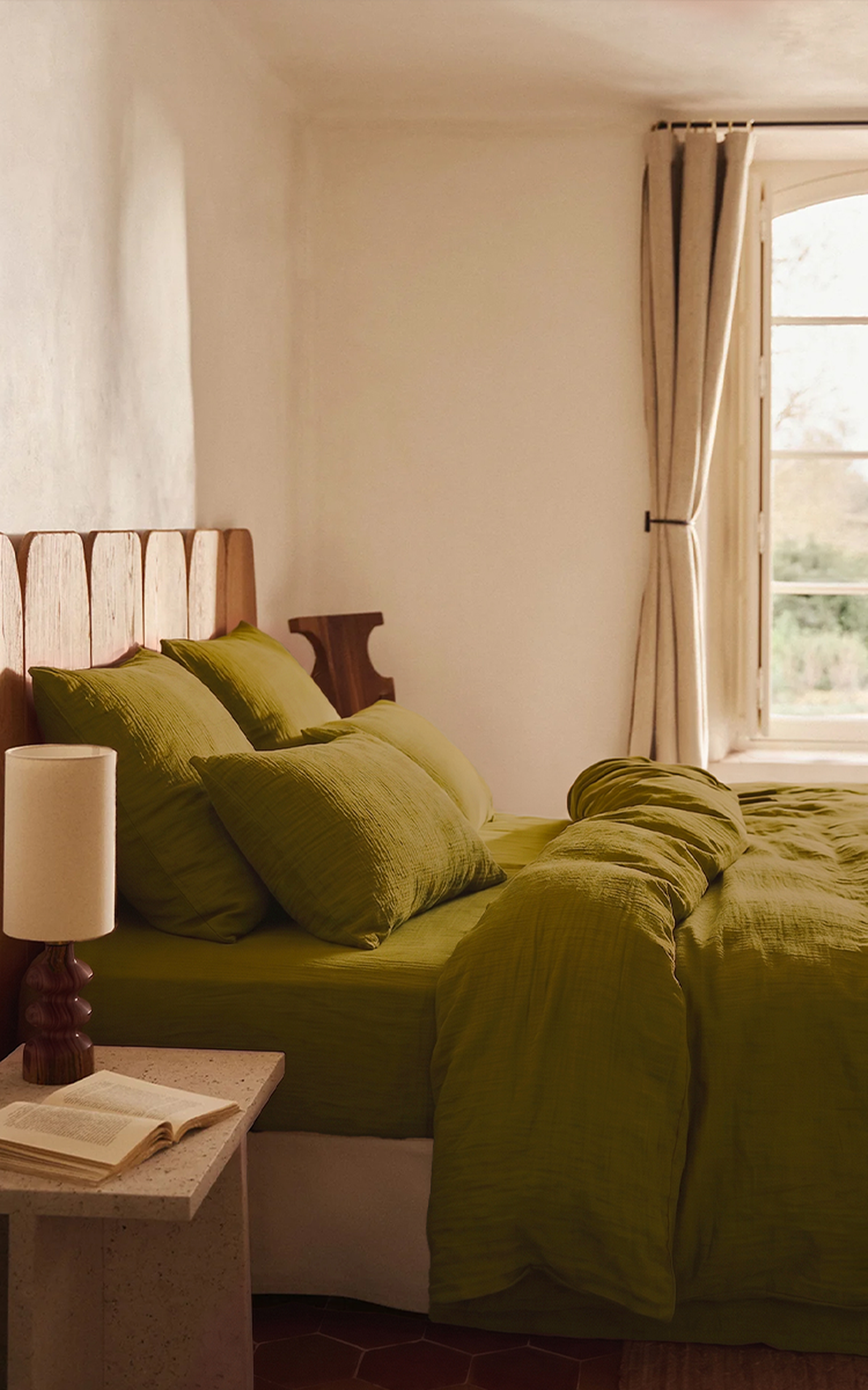 Une chambre douillette avec une tête de lit en bois, une parure de lit vert olive, la lumière du soleil qui pénètre à flots par la fenêtre, et une table de chevet sur laquelle sont posés une lampe et un livre ouvert.