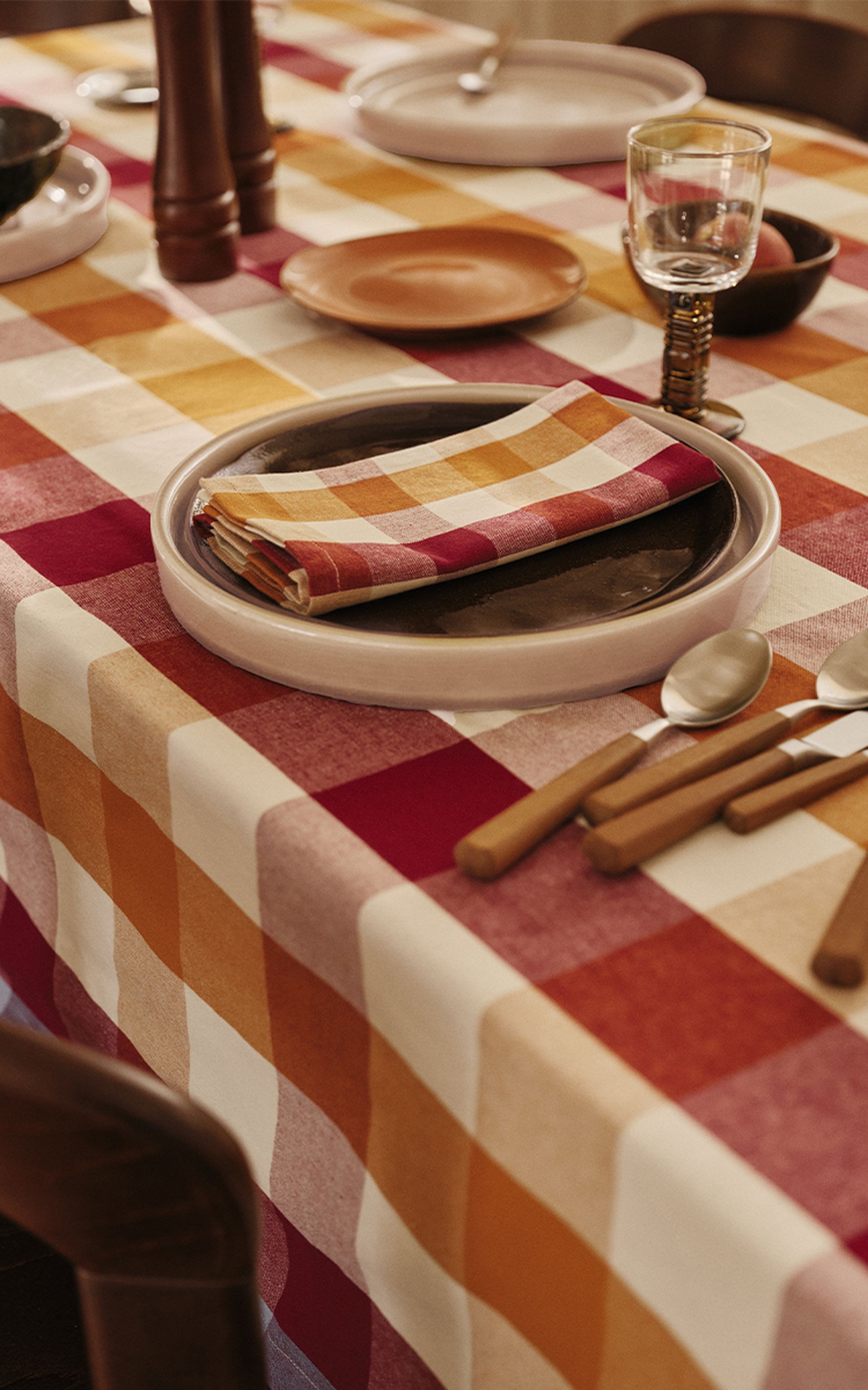 Une table dressée avec une nappe à carreaux, des assiettes empilées accompagnées d'une serviette assortie, des couverts, un verre et un moulin à poivre en bois