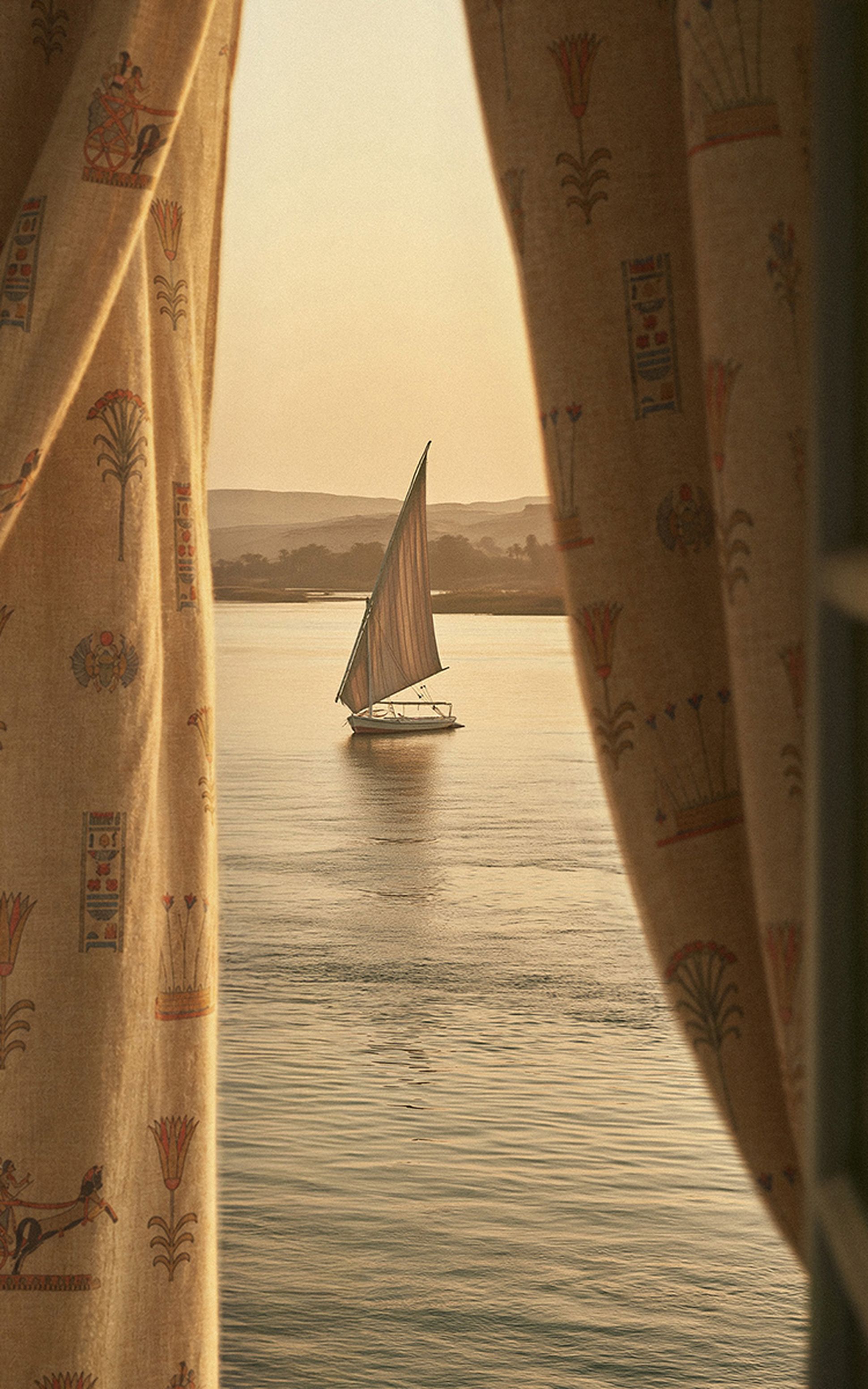 A sailboat with a single sail glides on calm water at sunset, framed by patterned curtains depicting ancient Egyptian motifs.