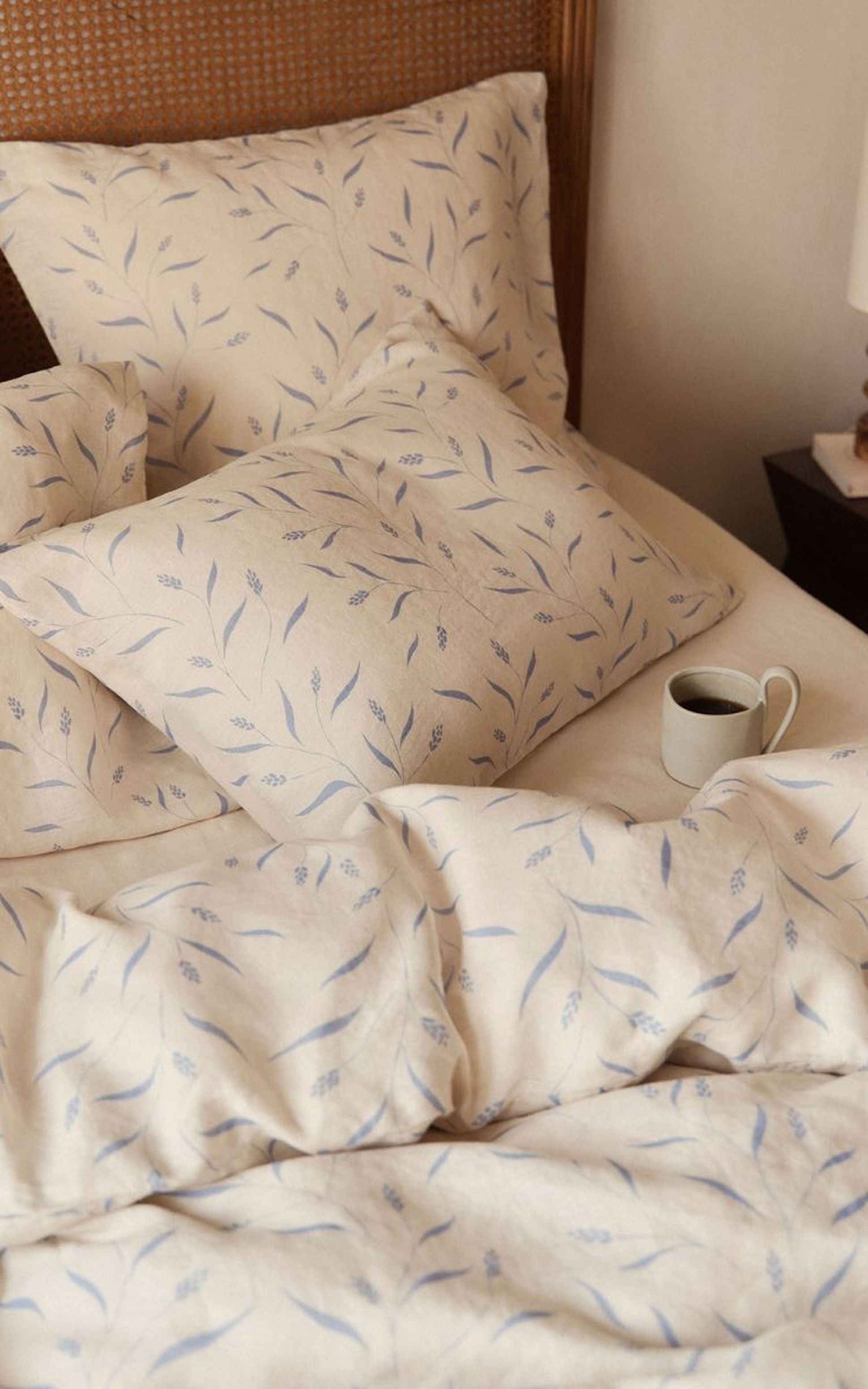 Une literie couleur crème à motifs de feuilles bleues sur un lit avec des oreillers ; une tasse de café est posée à côté de la literie.