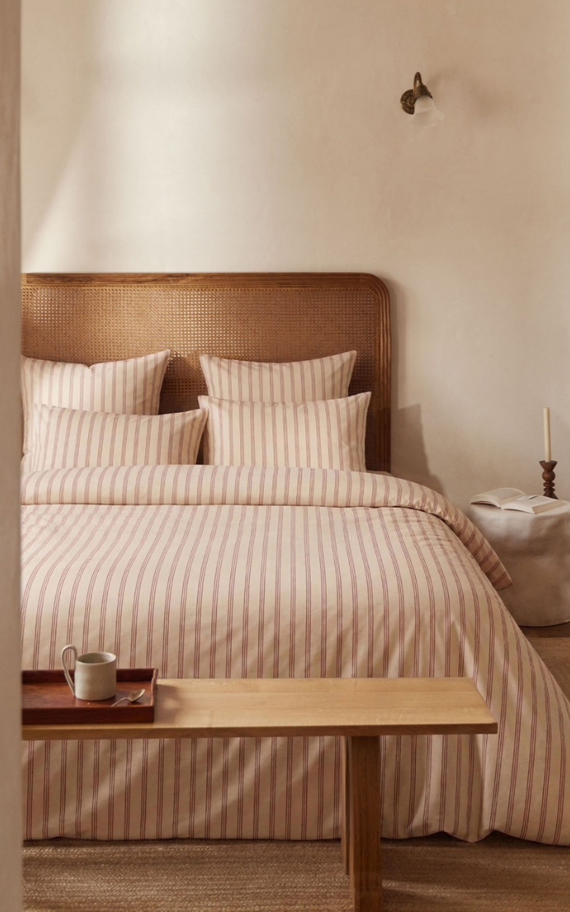 Une chambre lumineuse aux tons chaleureux, avec un lit soigneusement fait de draps beiges à fines rayures rouges. La tête de lit en cannage apporte une touche naturelle, tandis qu’une lampe élégante et un rideau clair complètent l’ambiance douce et apaisante.