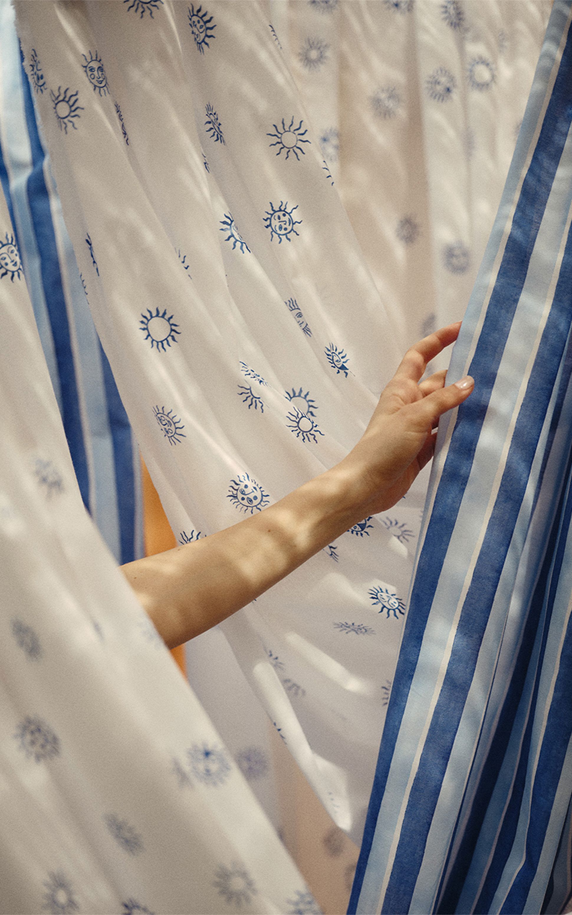 Une main effleure un rideau rayé bleu et blanc, entouré de rideaux blancs ornés de motifs de soleils bleus, créant ainsi une atmosphère sereine.