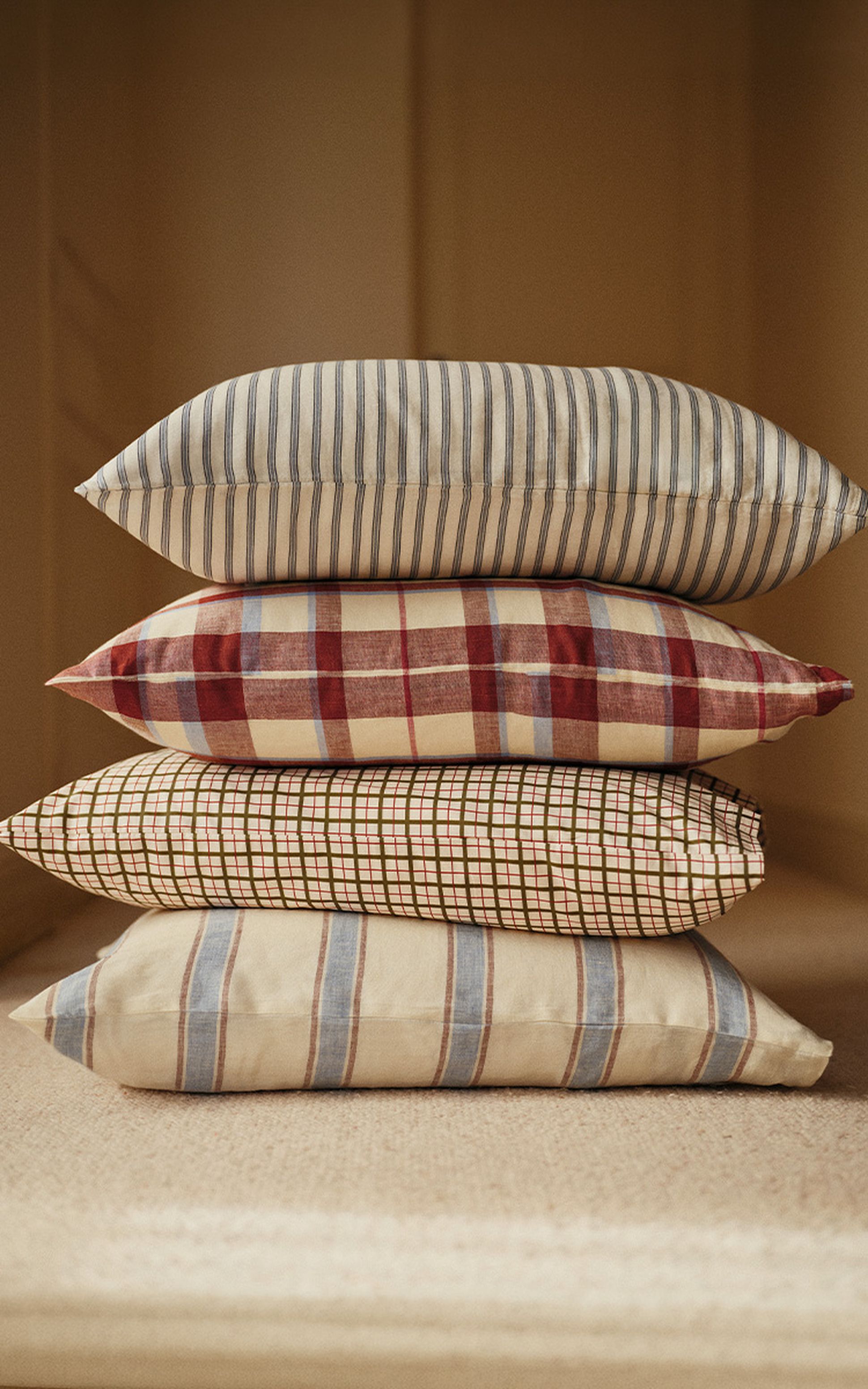 Une pile de quatre coussins décoratifs aux motifs variés à rayures et à carreaux, dans des tons de beige, de bleu et de rouge, sur un fond doux et neutre.