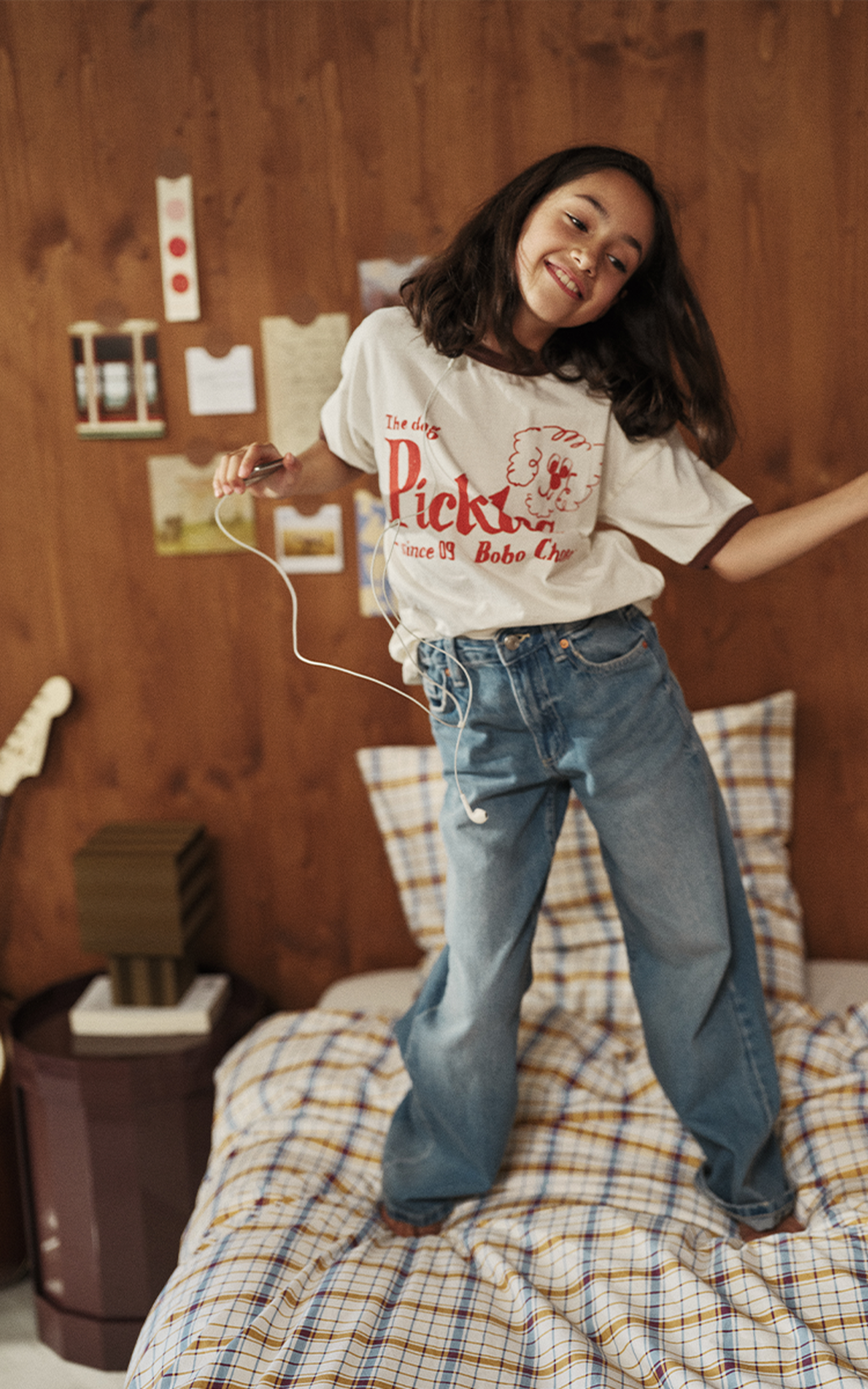 Une fille aux longs cheveux danse joyeusement sur un lit ; elle porte un t-shirt à motifs et un jean, ainsi qu'un casque audio, dans une chambre chaleureuse et décorée.