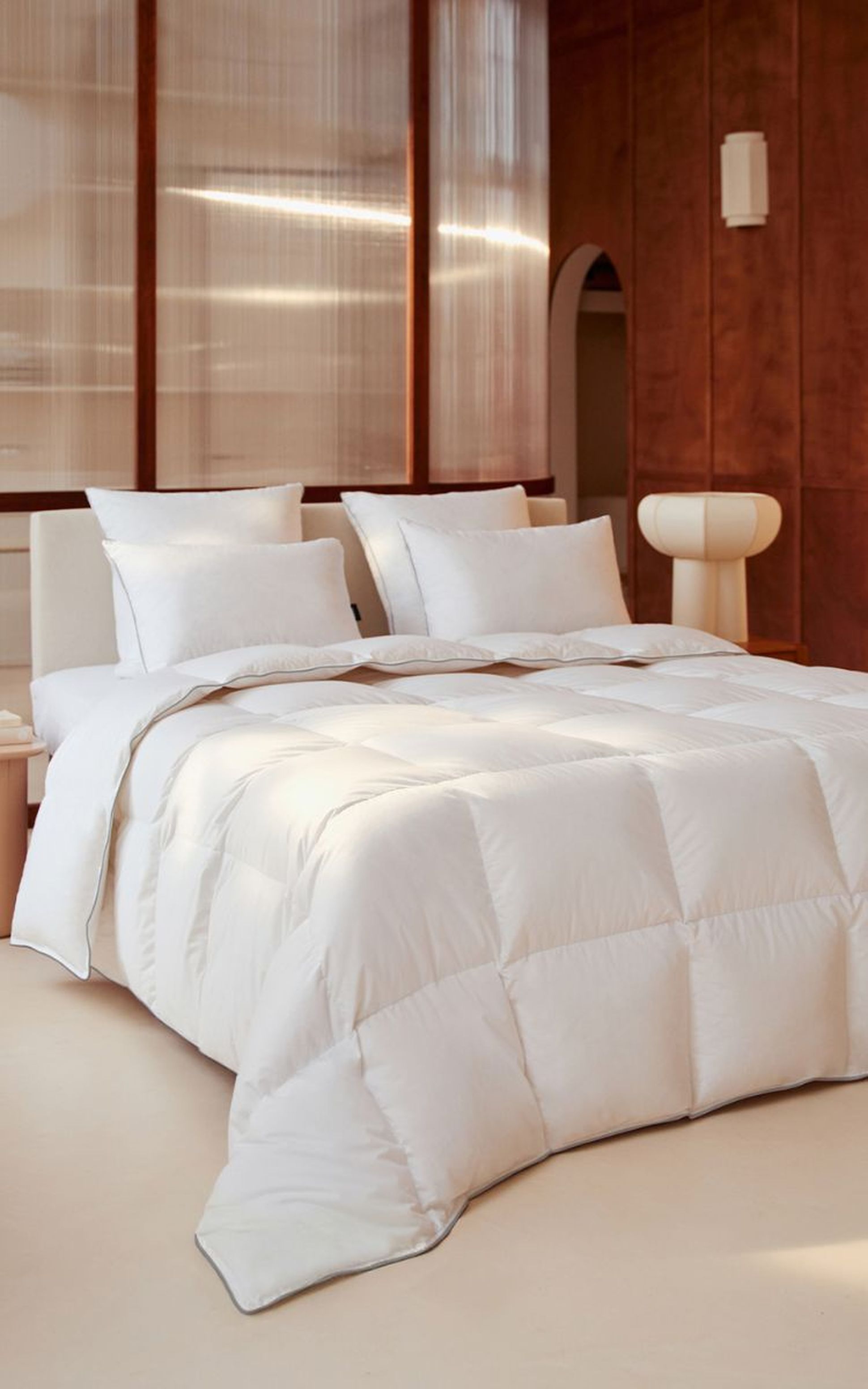 Une chambre moderne et chaleureuse dotée d'un lit avec une grande couette blanche et quatre oreillers blancs, d'un paravent aux tons neutres et d'un fauteuil beige élégant.