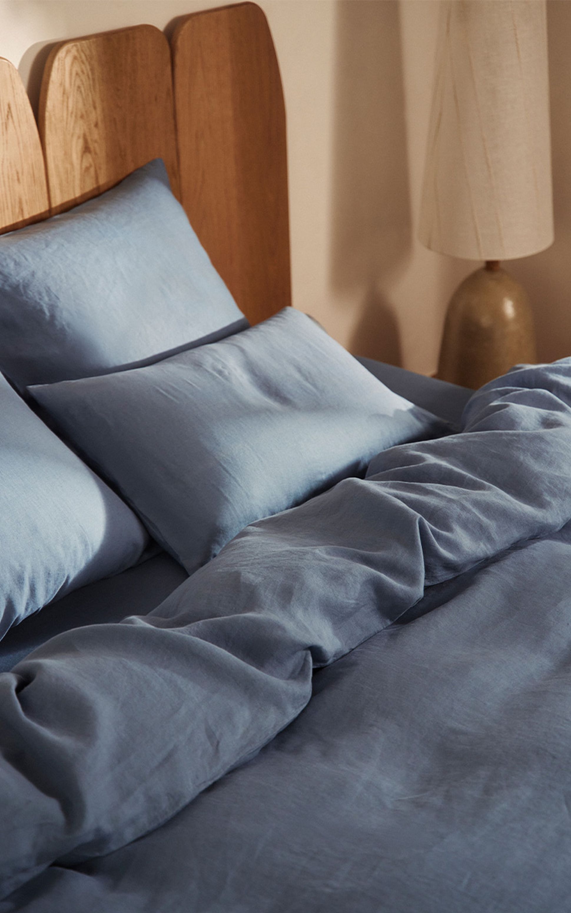 Lit douillet avec des oreillers et une couette bleu clair soigneusement disposés, une tête de lit en bois et une lampe beige dans une pièce faiblement éclairée.