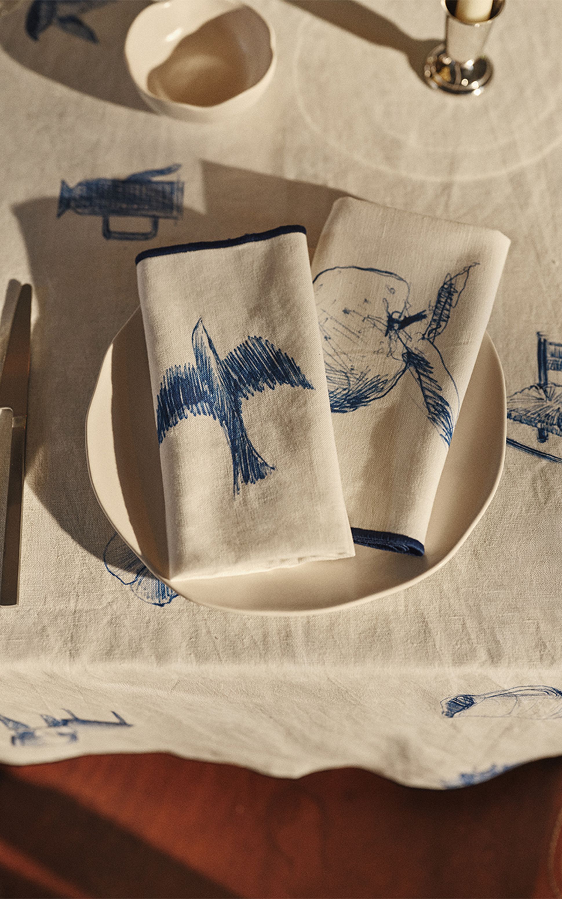 Deux serviettes blanches pliées, ornées de croquis d'oiseaux et de poissons bleus, posées sur une assiette blanche, le tout disposé sur une nappe aux motifs similaires, à côté d'un couteau et d'un bol.