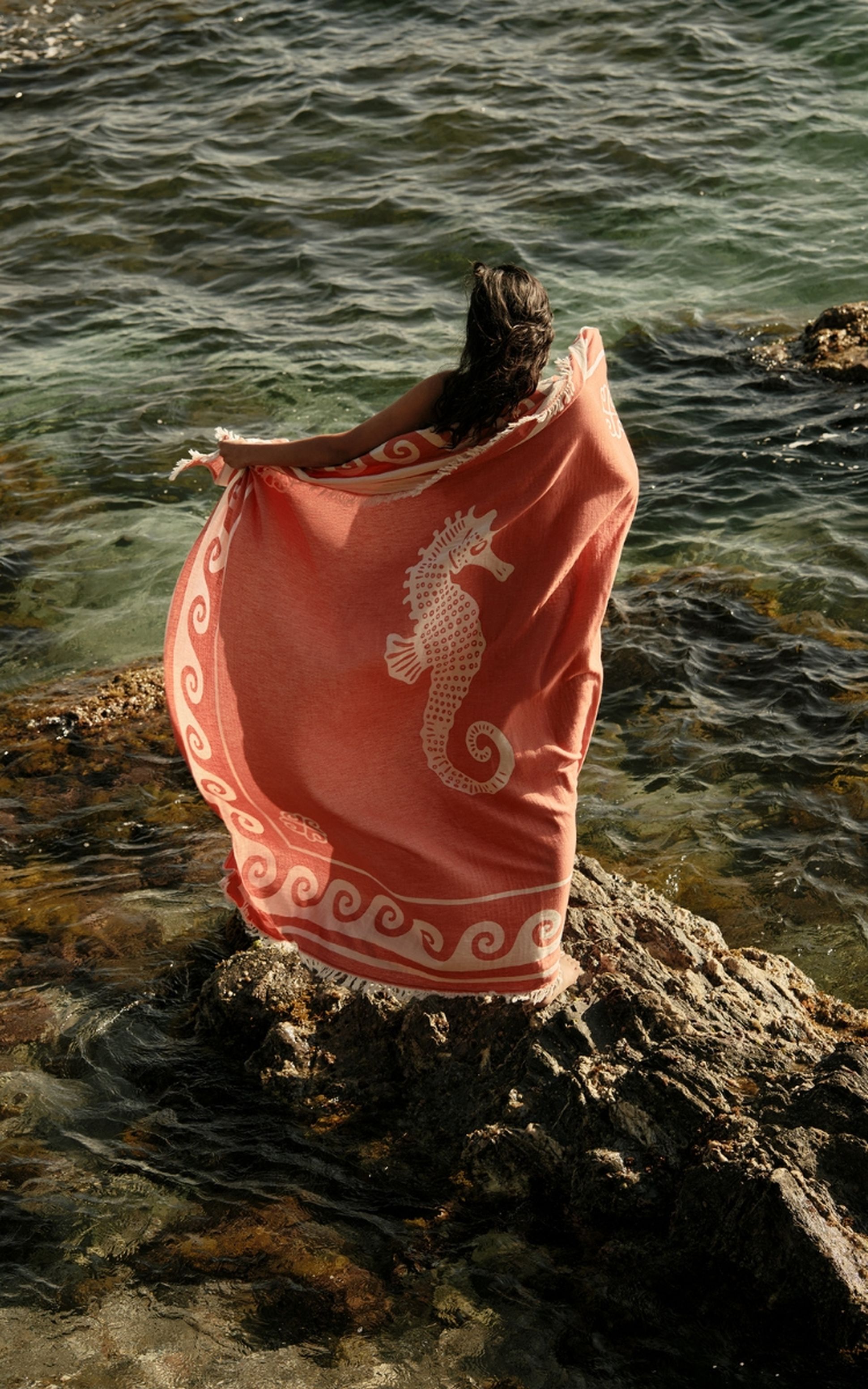 Une personne enveloppée dans une serviette rose ornée d'un motif d'hippocampe, debout sur des rochers au bord de la mer, contemplant l'eau limpide.