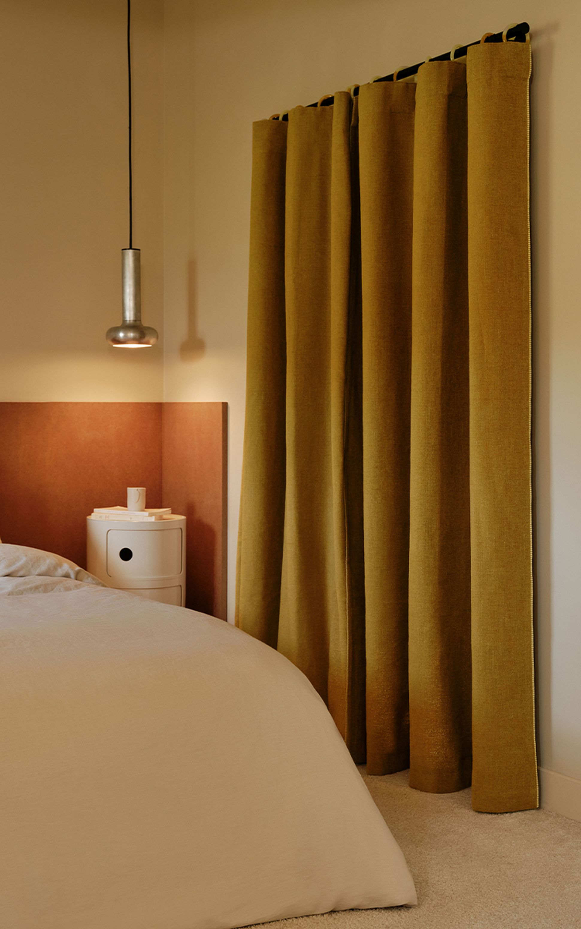 Cozy bedroom with mustard curtains, a pendant light, and a white nightstand beside a bed with a beige comforter.