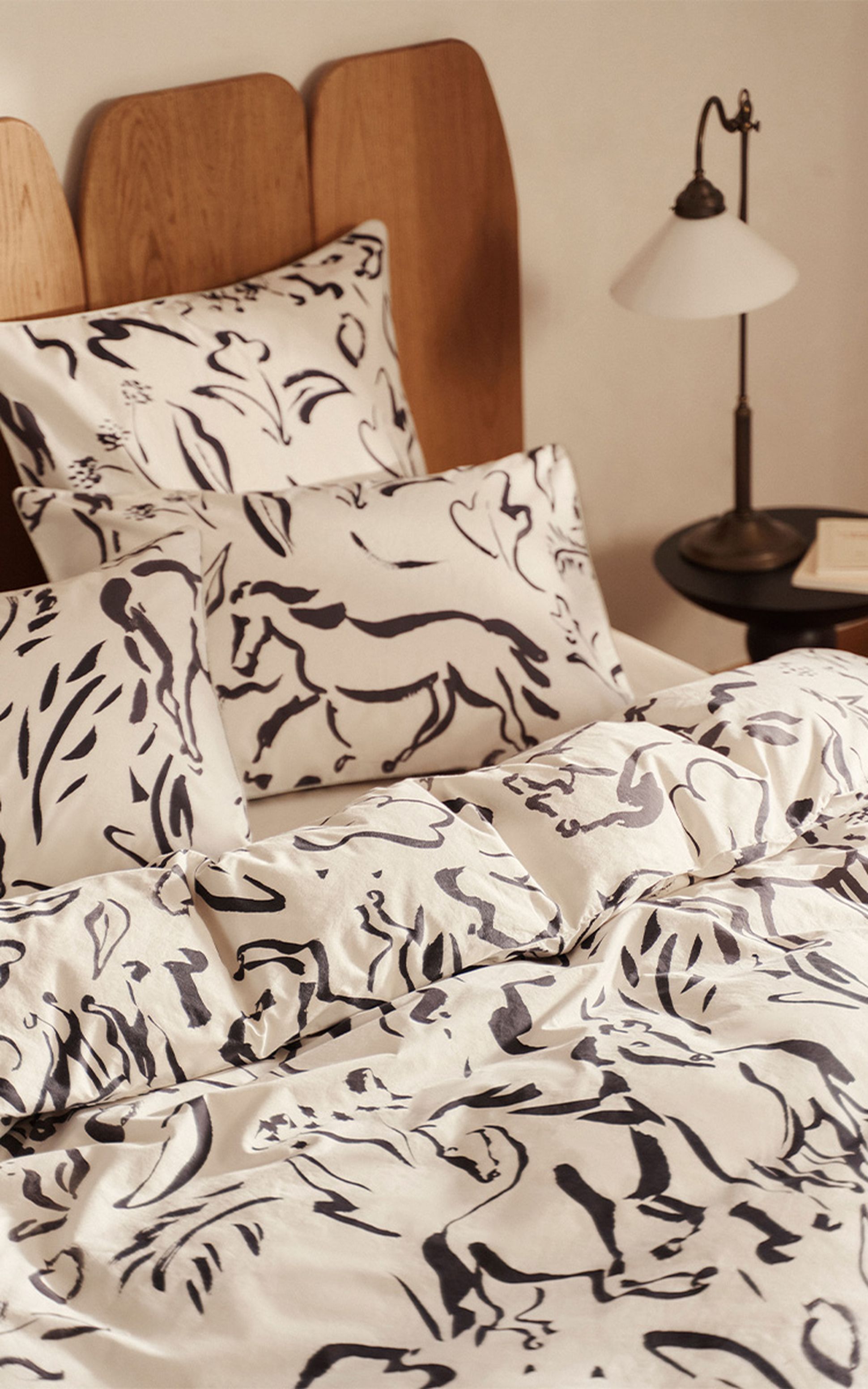 Ensemble de literie avec motifs abstraits de chevaux noirs sur des draps et des taies d'oreiller blancs, avec une tête de lit en bois et une table de chevet.