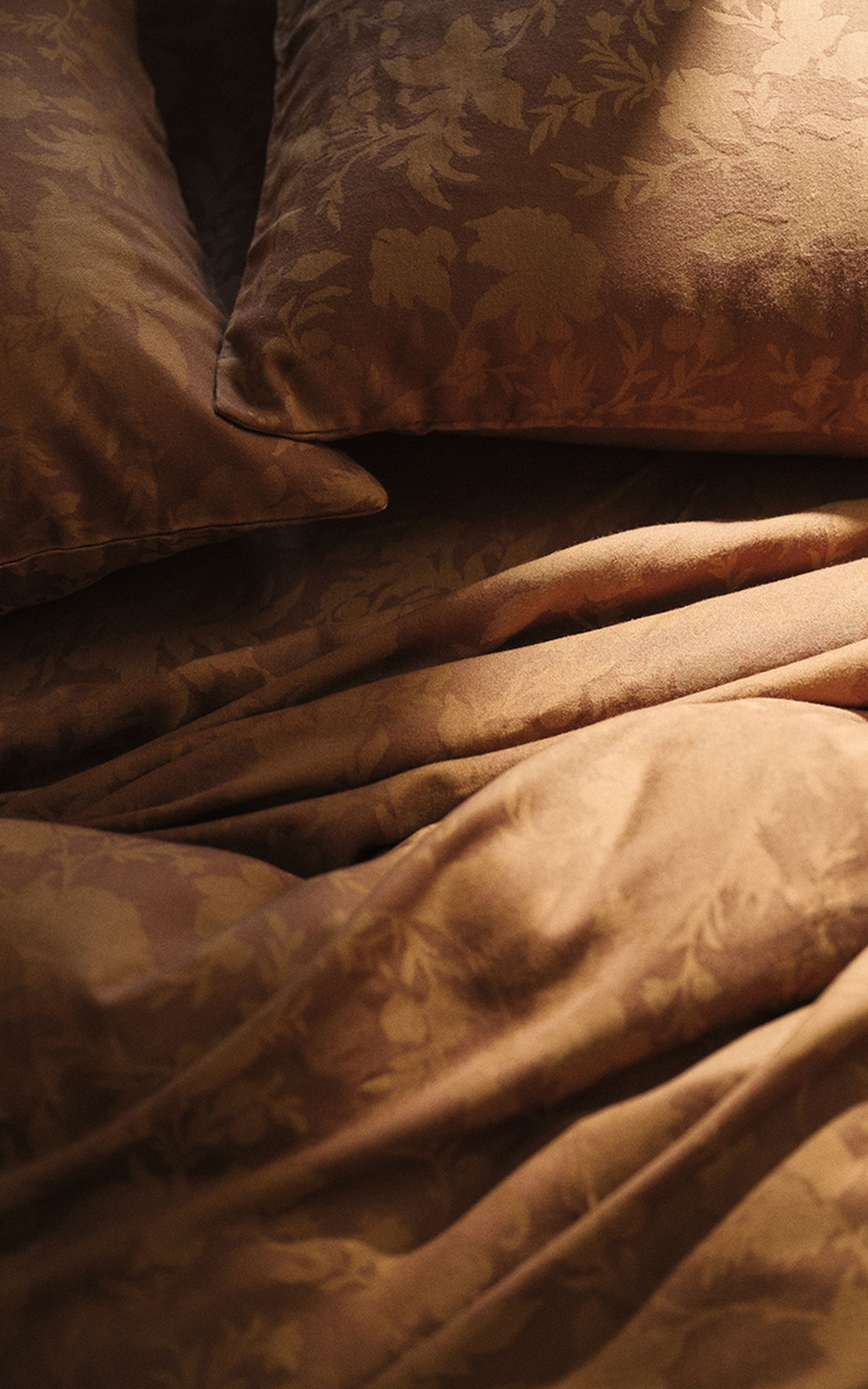 Gros plan sur un lit orné d'oreillers et de draps à motifs floraux marron, baigné d'une douce lumière naturelle qui crée une ambiance chaleureuse.