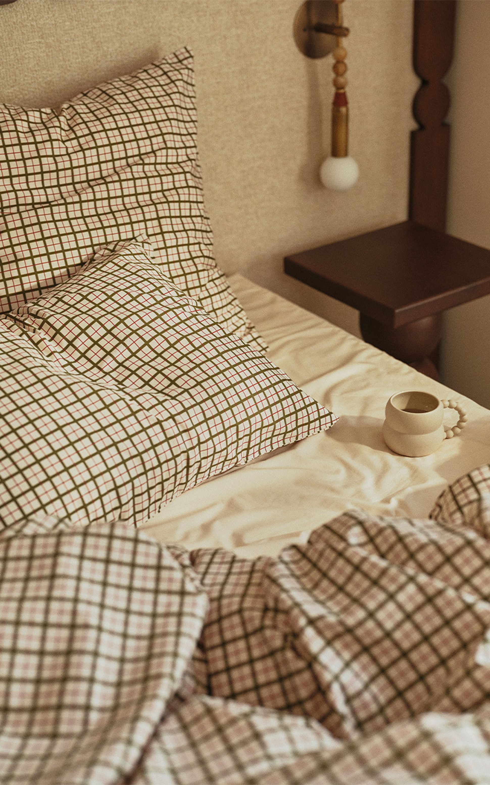 Un lit douillet avec des oreillers et une couette à carreaux, une table de chevet en bois et une tasse en céramique posée sur un drap blanc.