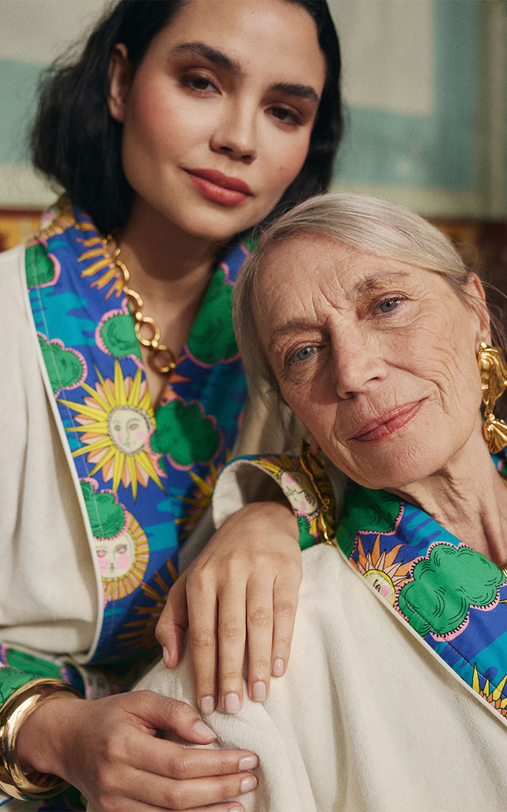 Deux femmes vêtues de tenues aux motifs colorés, l'une plus âgée aux cheveux gris et l'autre plus jeune aux cheveux foncés, posent côte à côte, dégageant une atmosphère chaleureuse.