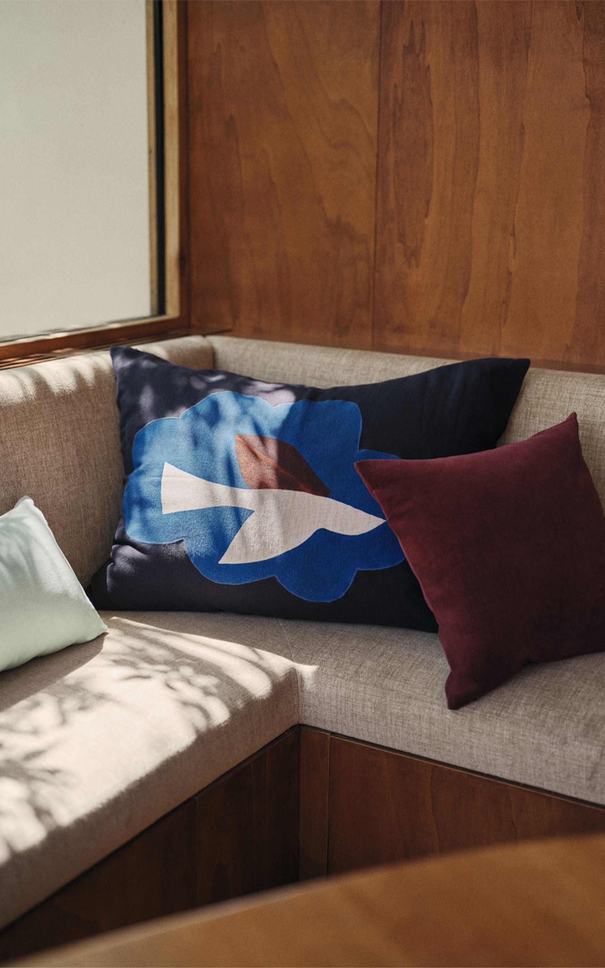 Corner sofa with beige cushions, featuring three pillows: one with a blue abstract design, a white one, and a dark red one, against a wooden wall.