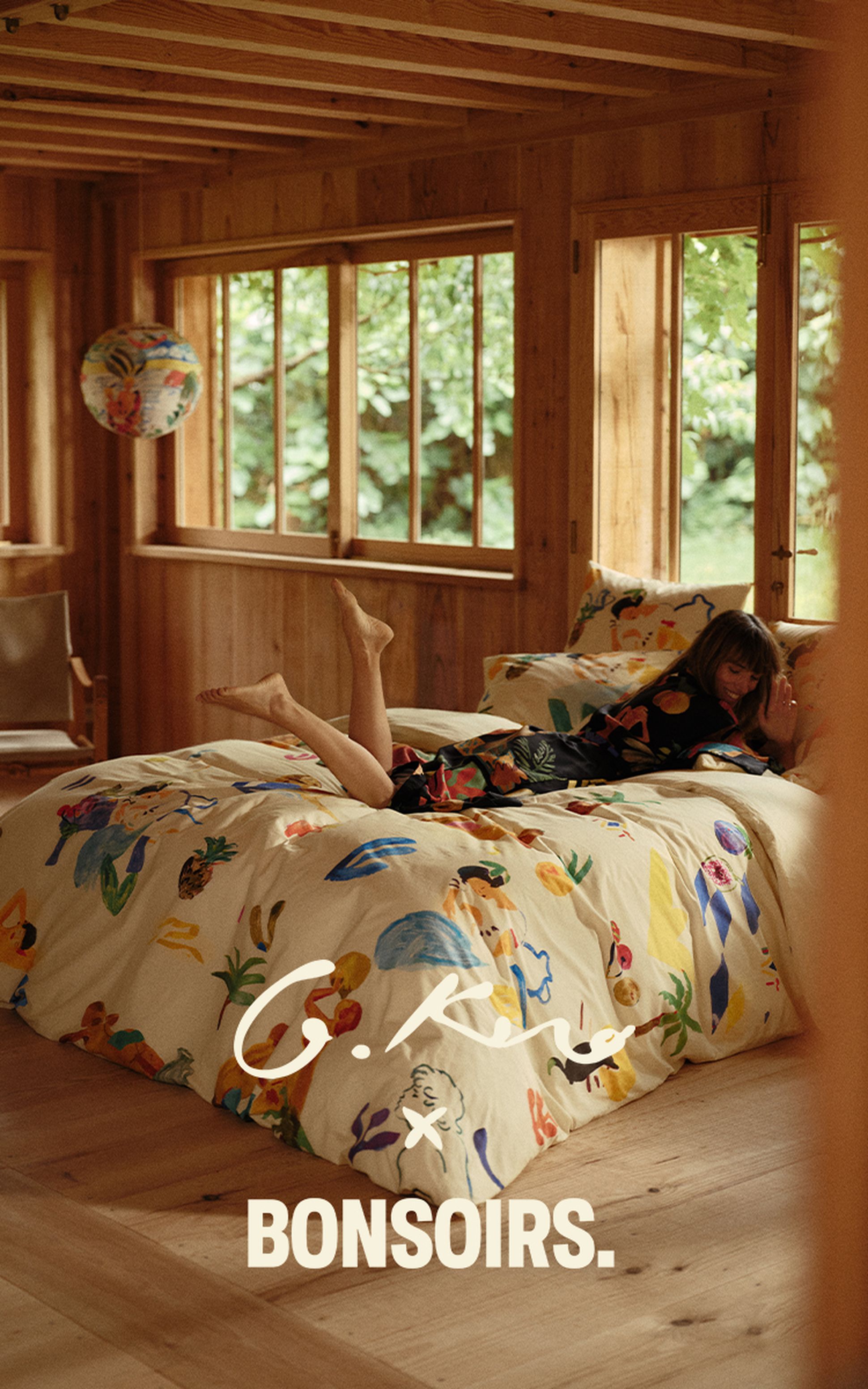 Une femme allongée sur un lit douillet aux draps colorés, dans une chambre aux murs en bois, avec de grandes fenêtres donnant sur la verdure ; on peut lire « G. Kero x Bonsoirs ».