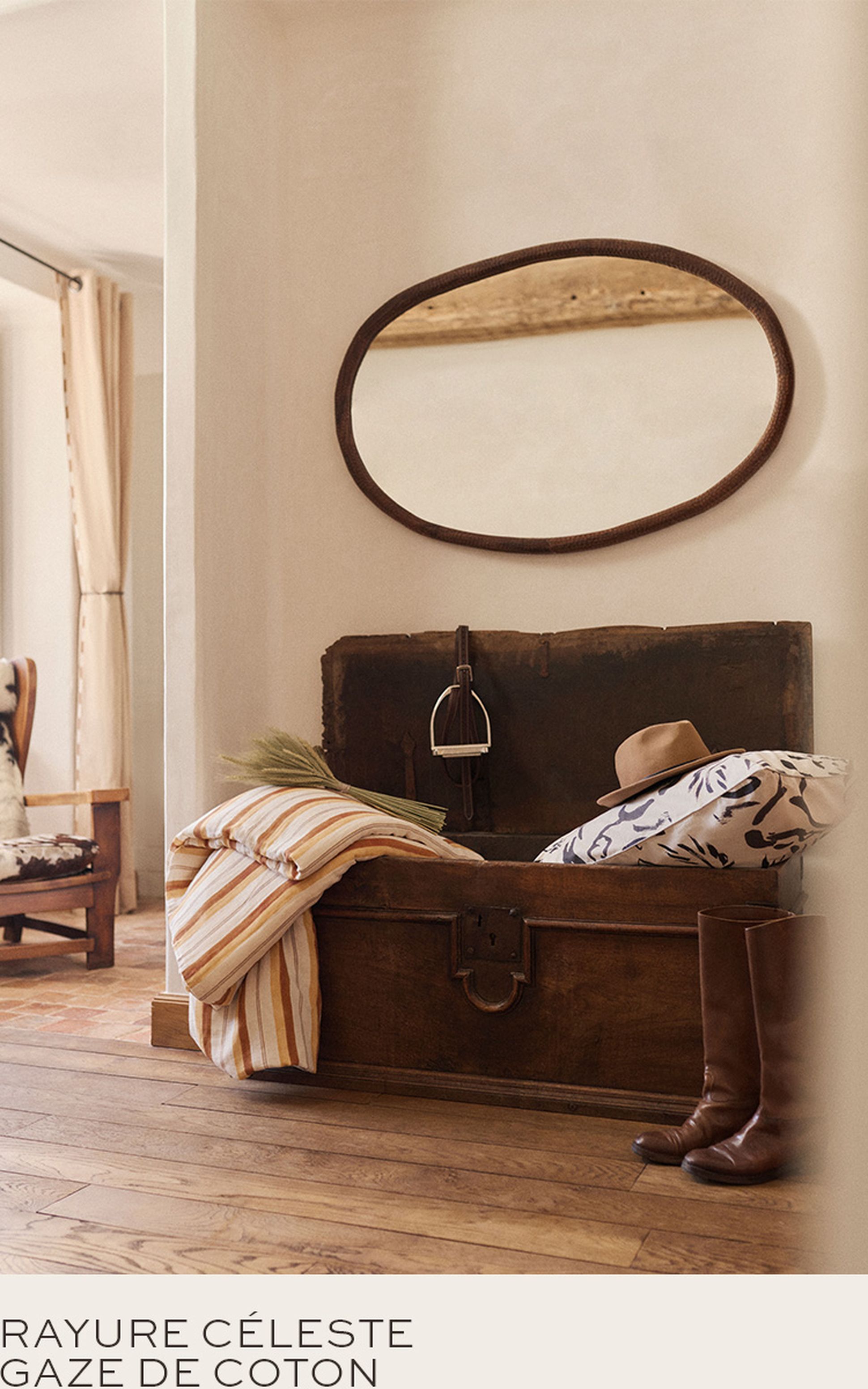 Une malle rustique en bois contenant des coussins, un chapeau et des bottes est placée sous un miroir ovale dans une pièce chaleureusement éclairée avec un sol en bois.