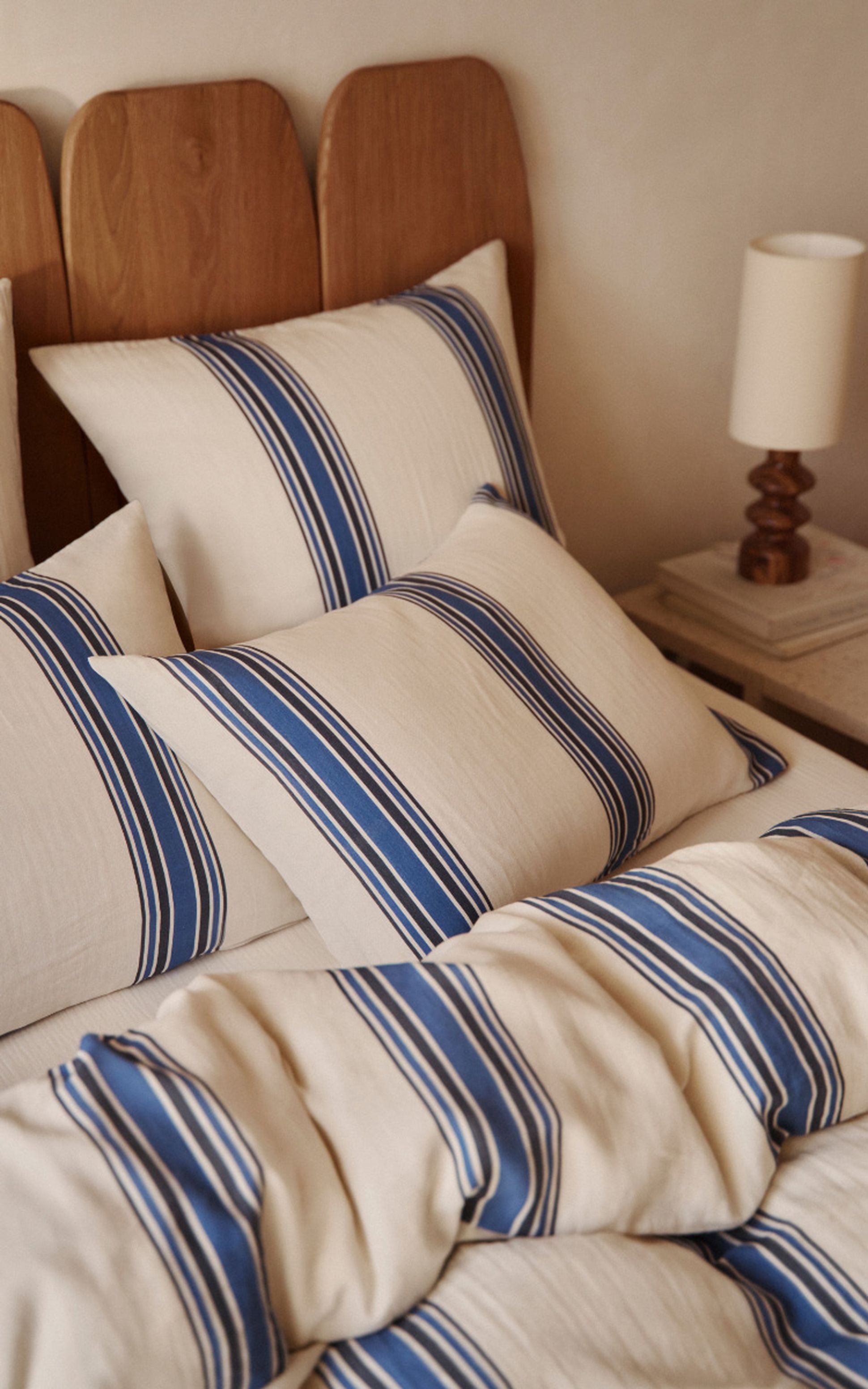 Un lit soigneusement fait, orné d'oreillers et d'une couette à rayures crème et bleues, avec une tête de lit en bois et une lampe posée sur une table de chevet.
