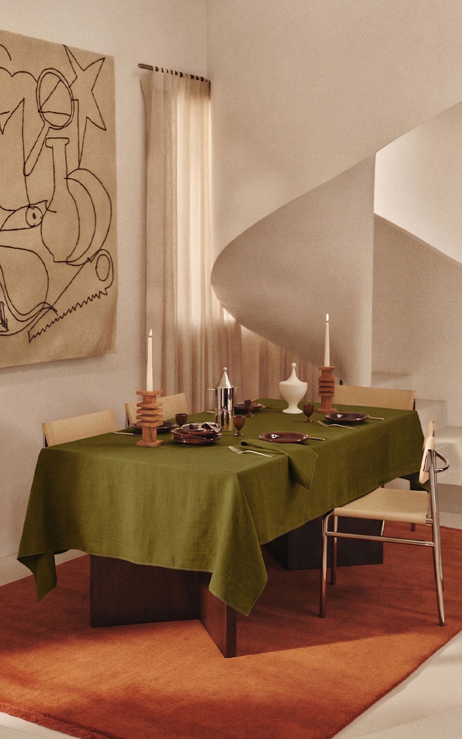 Une salle à manger moderne avec une nappe verte, décorée de bougies, de vaisselle et d'un pichet en argent. Les murs beiges sont ornés d'œuvres d'art abstrait et d'un escalier en colimaçon.
