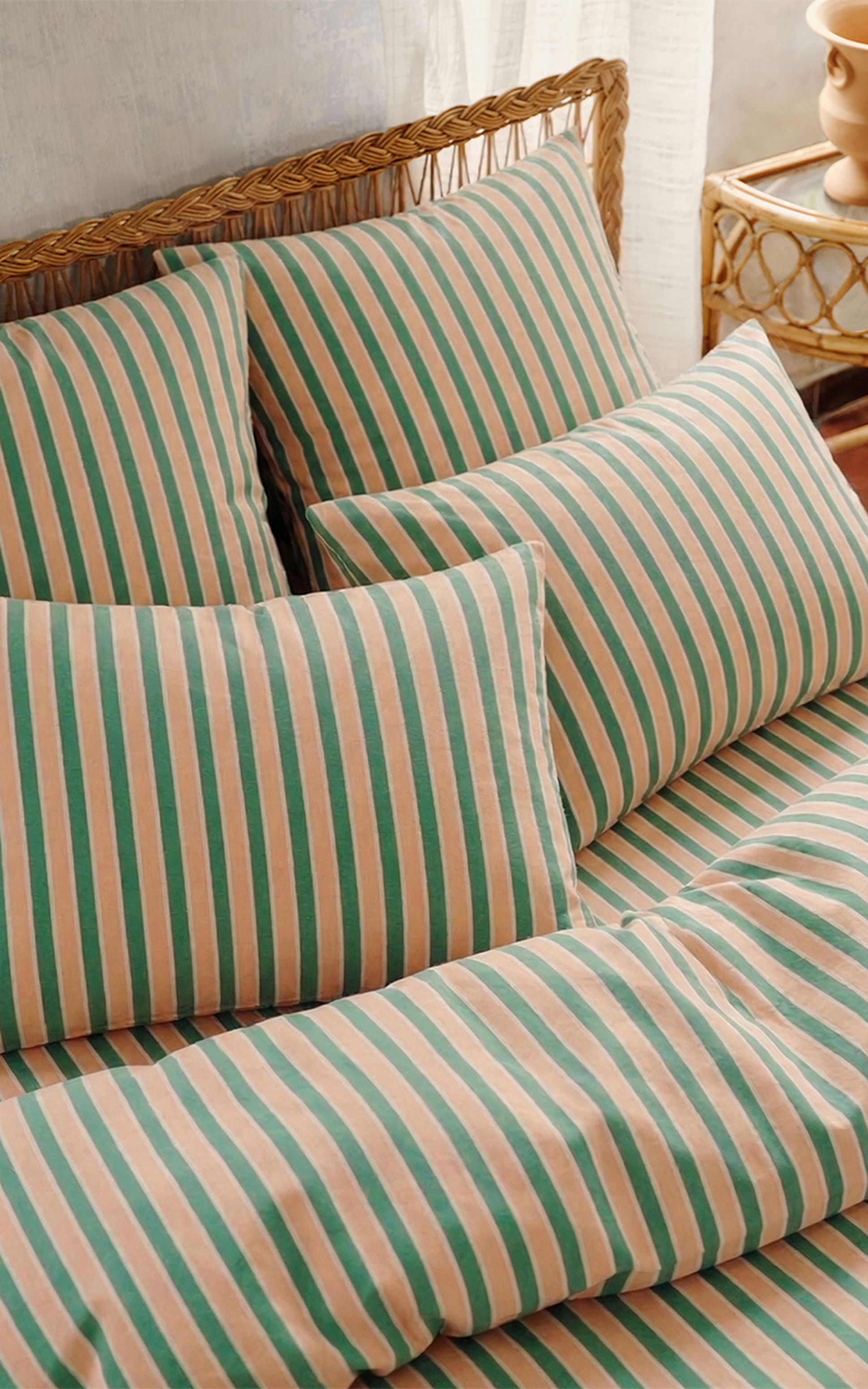 Des coussins et une parure de lit rayés, dans des tons vert pastel et pêche, sur un lit en rotin accompagné d'une petite table de chevet.