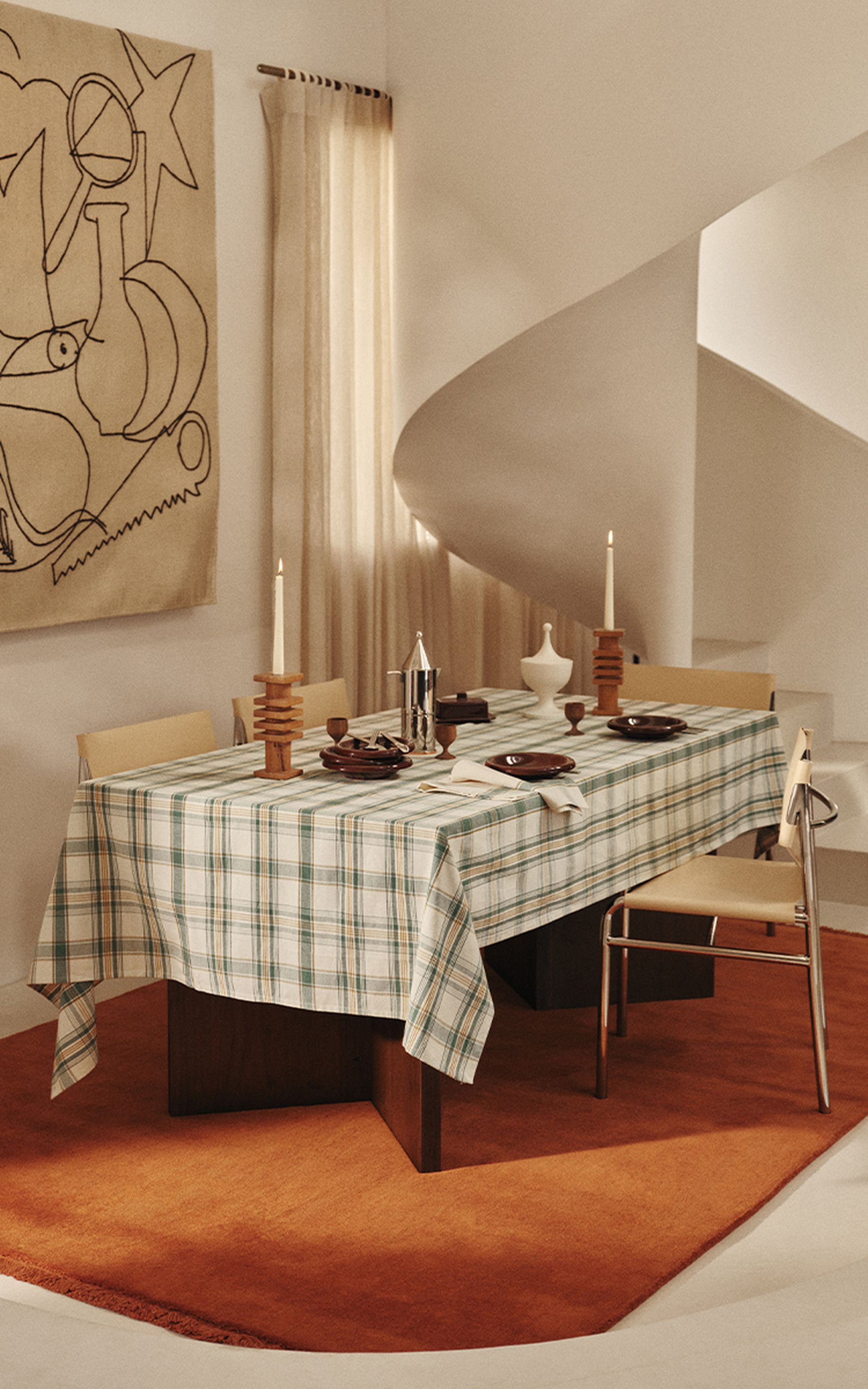 A dining room with a checkered tablecloth, set for a meal with plates and cutlery, surrounded by chairs, on an orange rug, near a curved staircase.