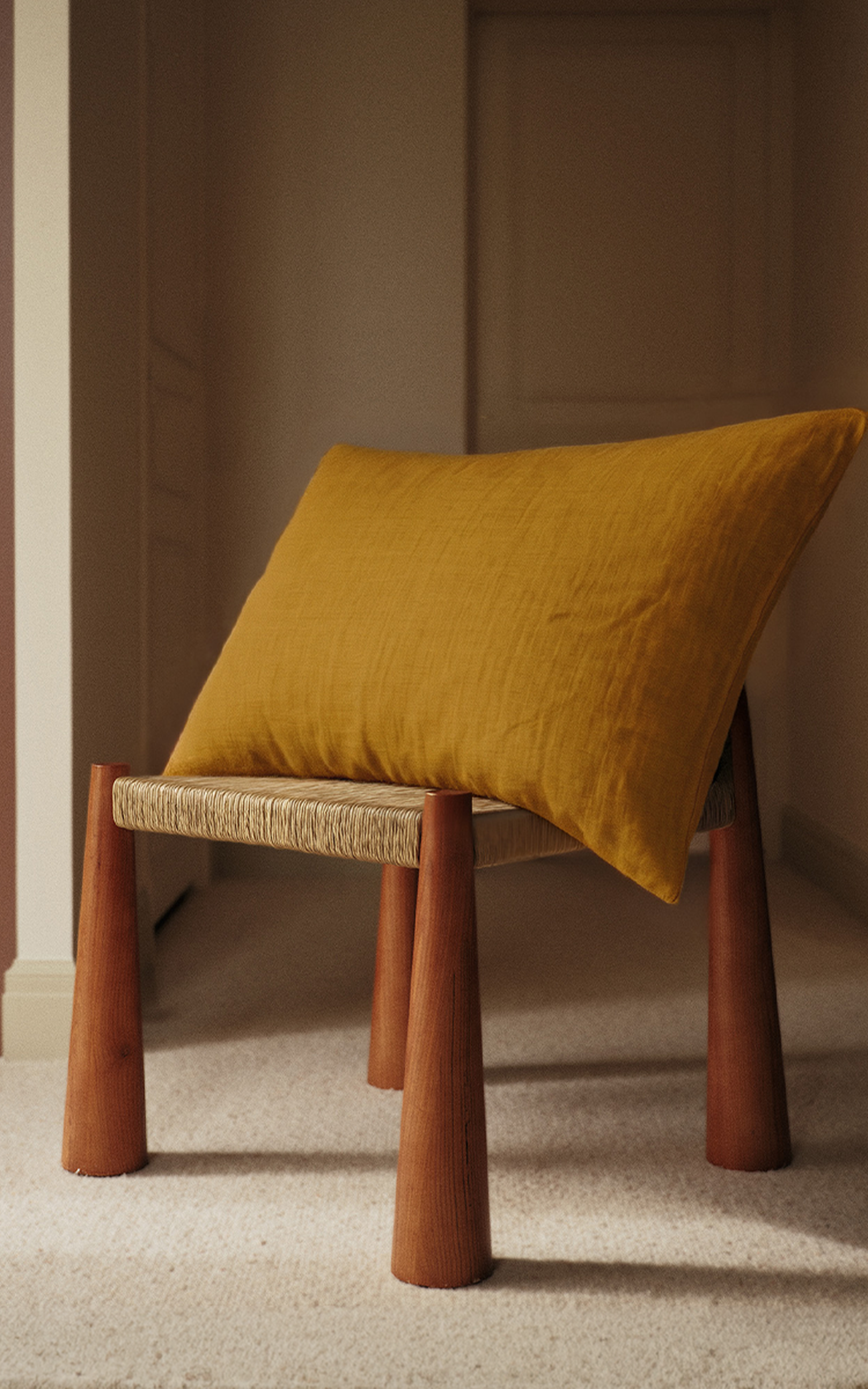 Un tabouret aux pieds en bois et à l'assise tressée est orné d'un coussin jaune moutarde, placé dans une pièce baignée d'une lumière tamisée.