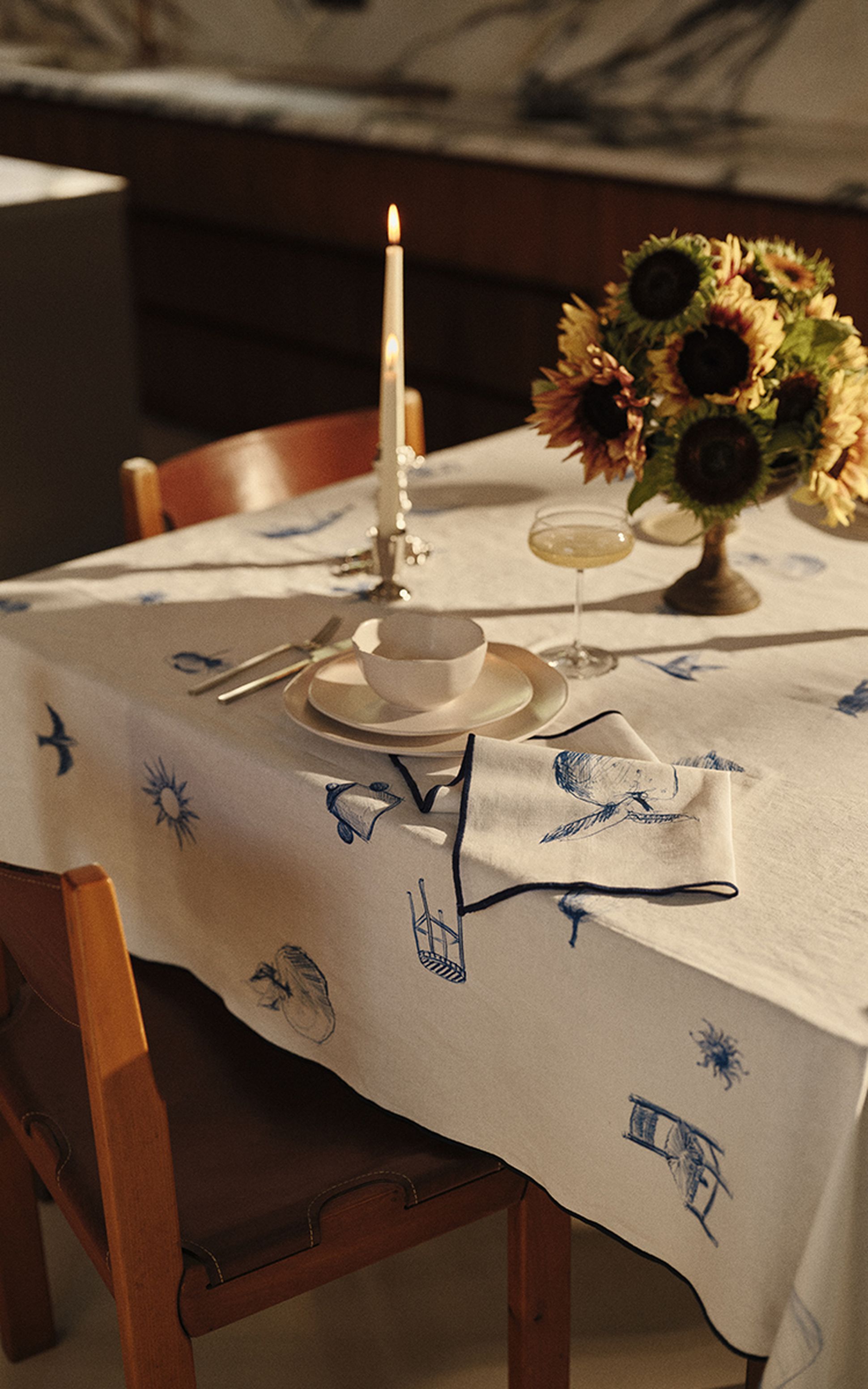Une table élégamment dressée, avec une nappe blanche ornée de motifs bleus, une bougie allumée, un centre de table composé de tournesols, de la vaisselle et des verres.