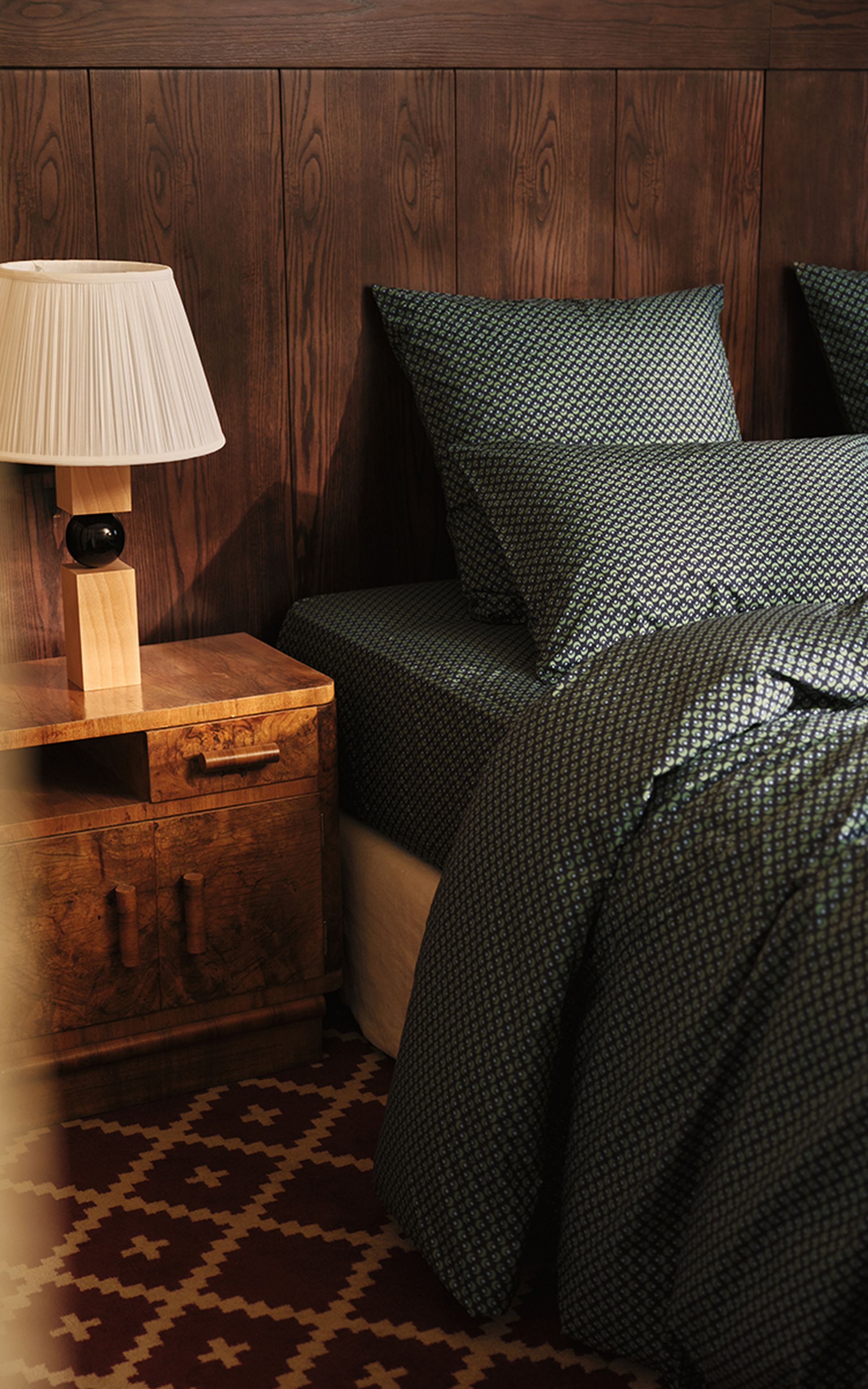 Une chambre à coucher chaleureuse, avec une table de chevet en bois, une lampe et un lit orné d'une parure à motifs, le tout devant un mur lambrissé.