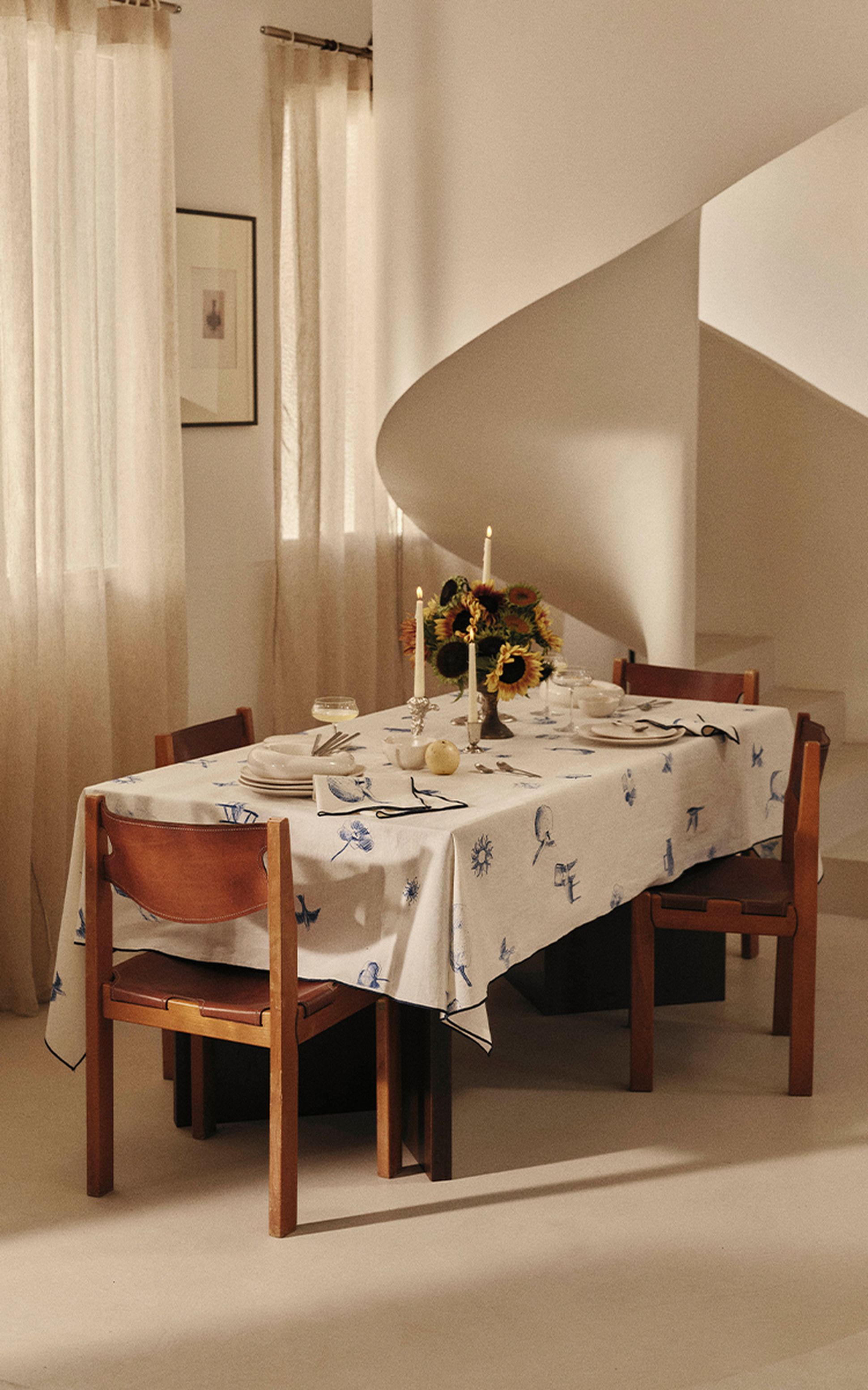 Une table à manger dressée pour quatre personnes, avec une nappe à motifs, des bougies et des tournesols, à côté d'un escalier en colimaçon et de rideaux légers et vaporeux.