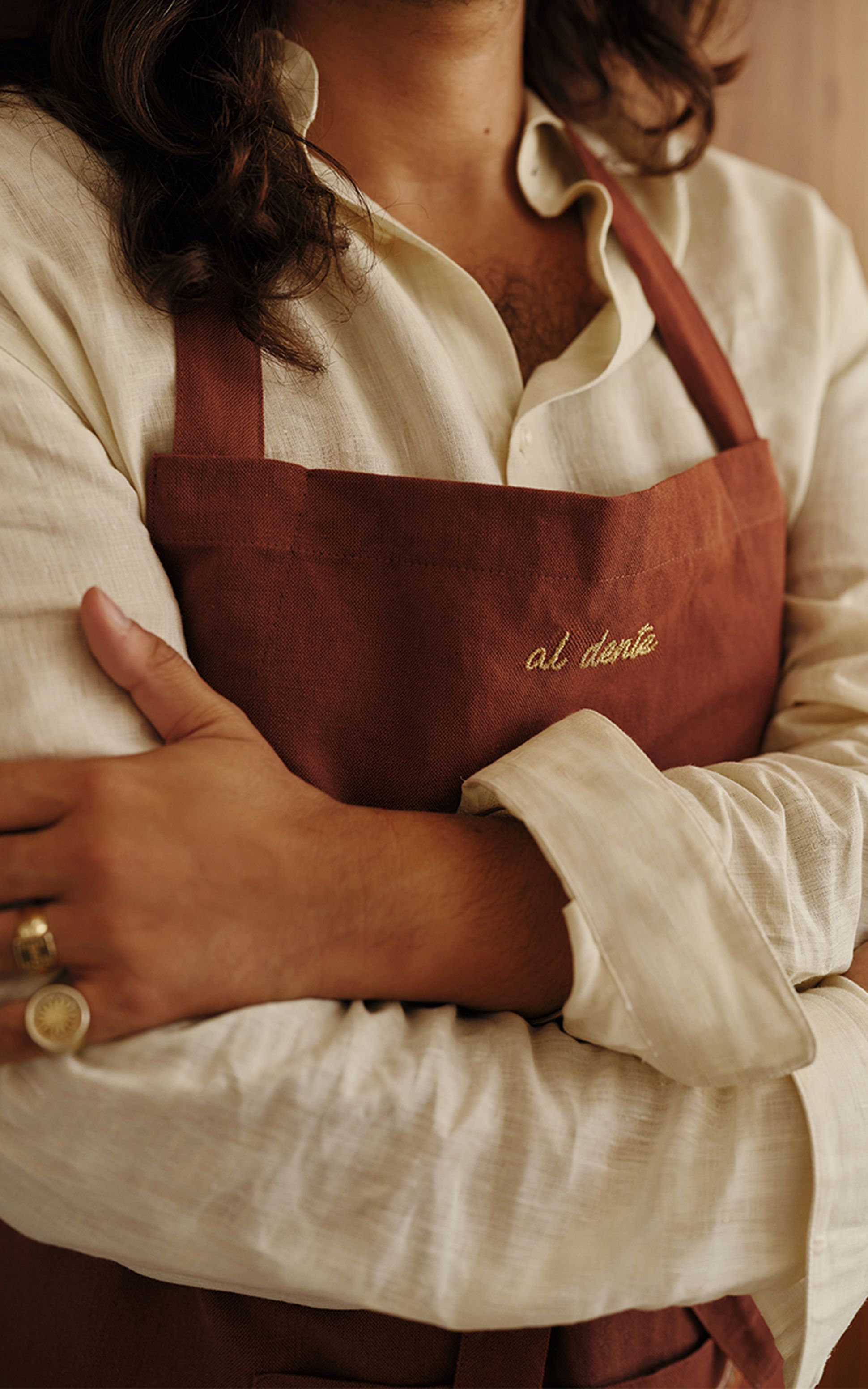 Personne vêtue d'une chemise beige et d'un tablier marron sur lequel est brodé « al dente », les bras croisés, avec des bagues aux doigts.