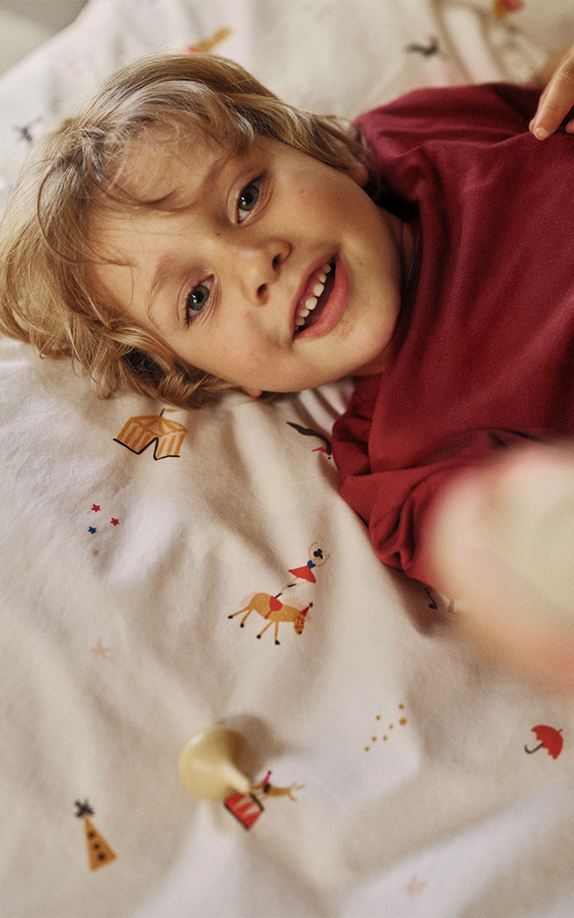 Un enfant vêtu d'un t-shirt rouge sourit en étant allongé sur un lit. La literie est ornée d'illustrations colorées et ludiques, telles qu'un cheval et des étoiles.