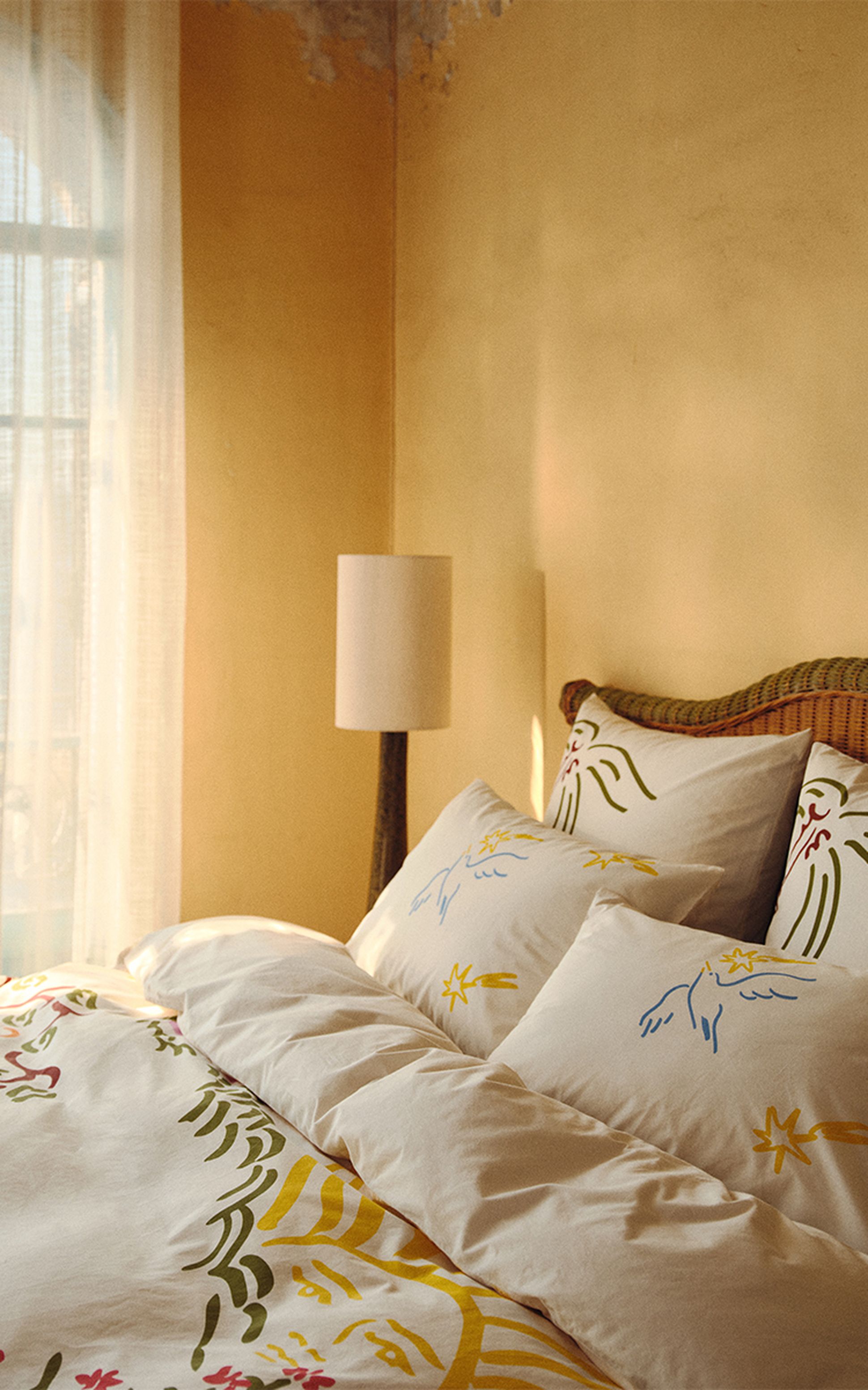 Une chambre baignée de soleil, avec une parure de lit blanche ornée de motifs abstraits colorés, une tête de lit en osier et un grand lampadaire placé près d'une fenêtre aux rideaux vaporeux.