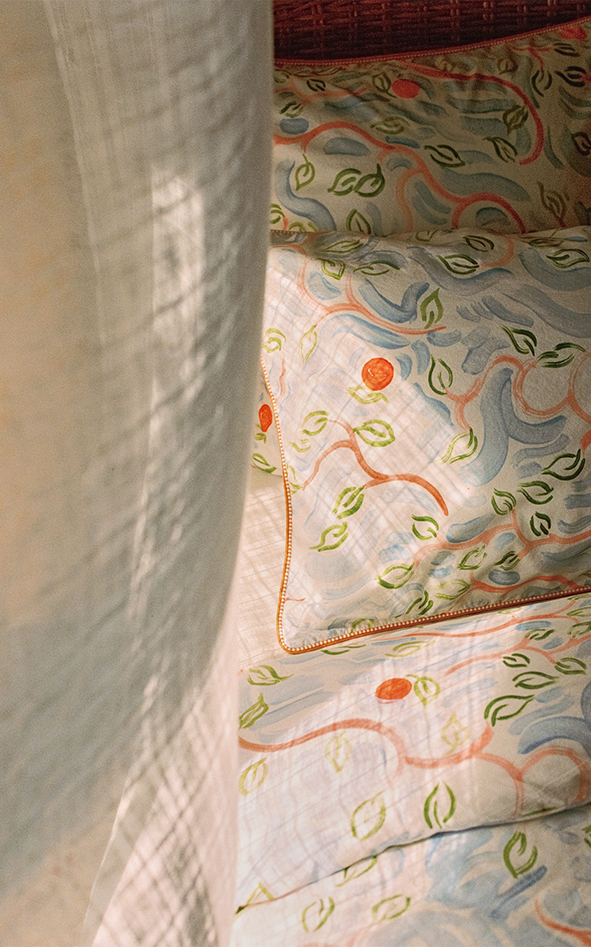 Gros plan sur un lit recouvert de draps colorés à motifs, représentant des cercles orange et des feuilles vertes, partiellement masqués par un rideau léger et vaporeux.