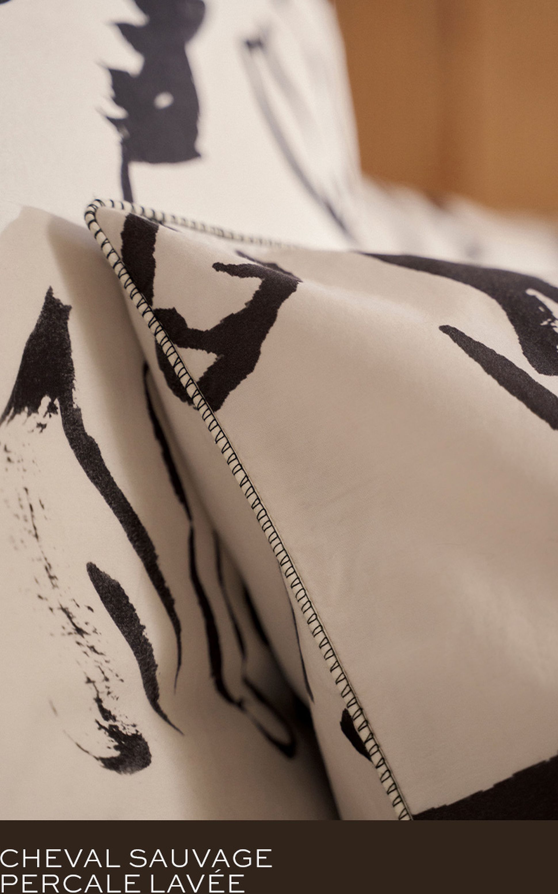 Gros plan sur un tissu beige avec des motifs abstraits noirs et des bords cousus. Texte : « Cheval Sauvage Percale Lavée » en blanc sur fond sombre.