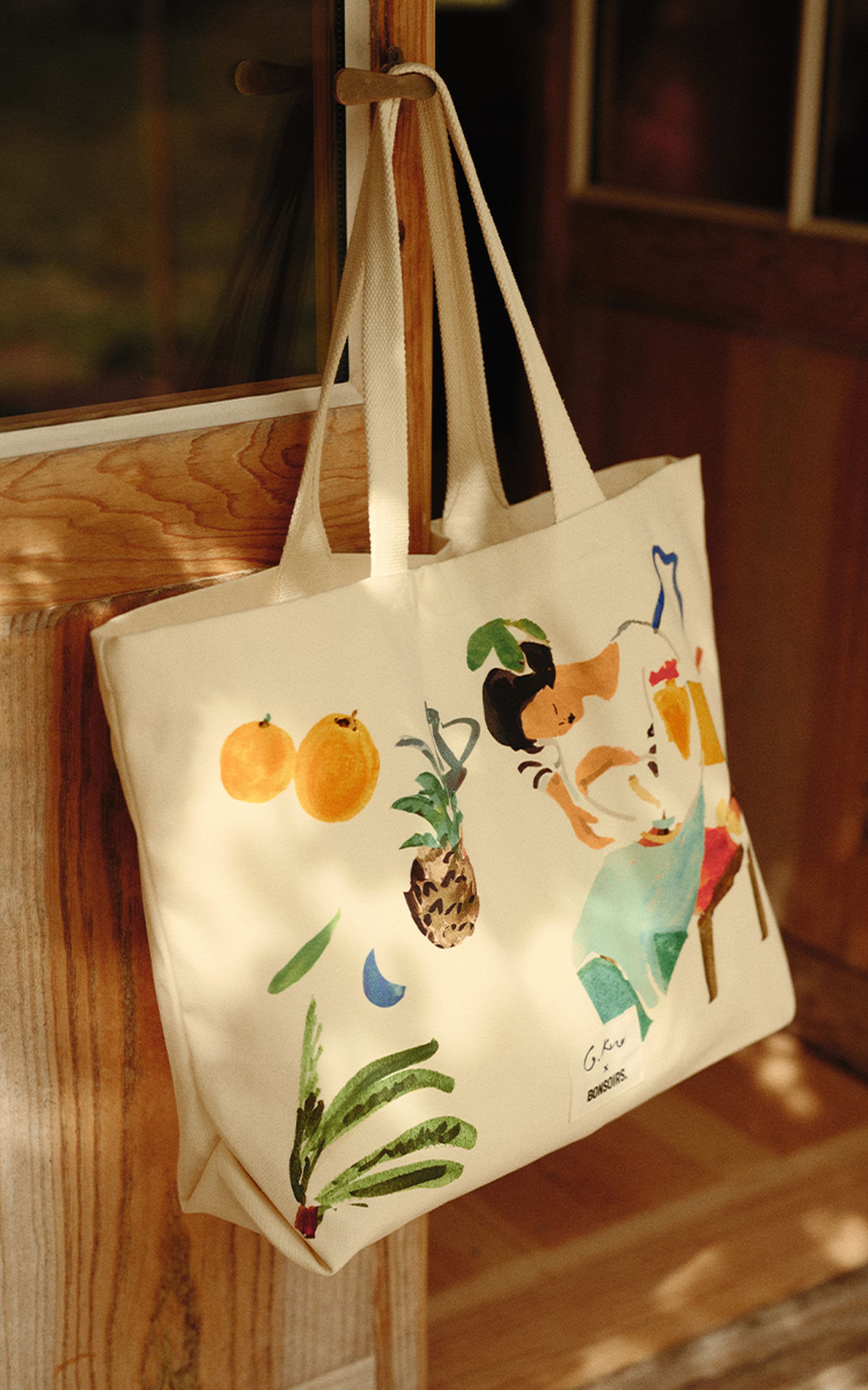 Sac fourre-tout en toile orné d'une illustration colorée représentant des fruits et une femme, suspendu à une poignée de porte sous un éclairage chaleureux.
