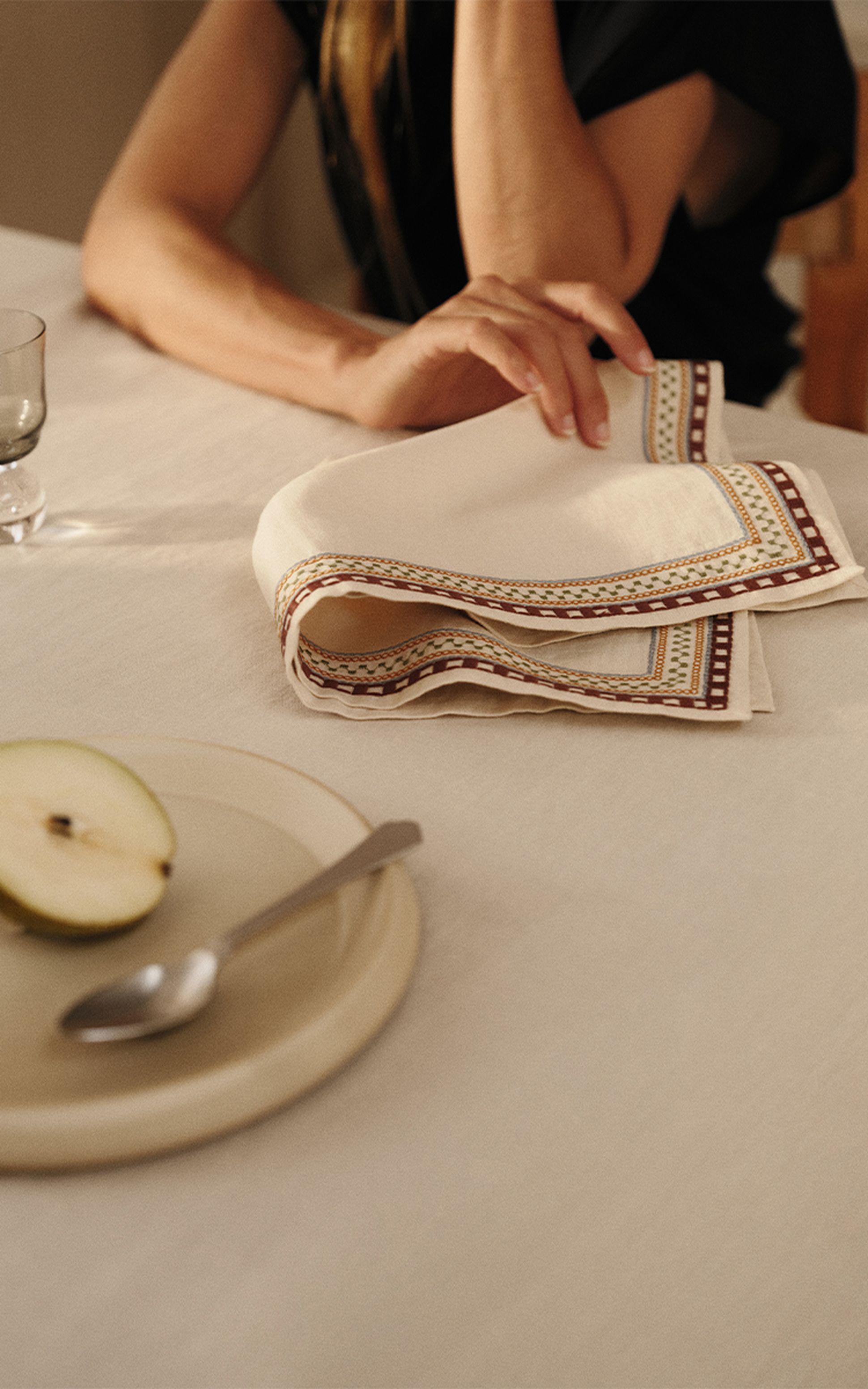 Une personne aux cheveux longs est en train de plier une serviette décorative sur une table, à côté d'une assiette contenant une demi-poire et une cuillère.
