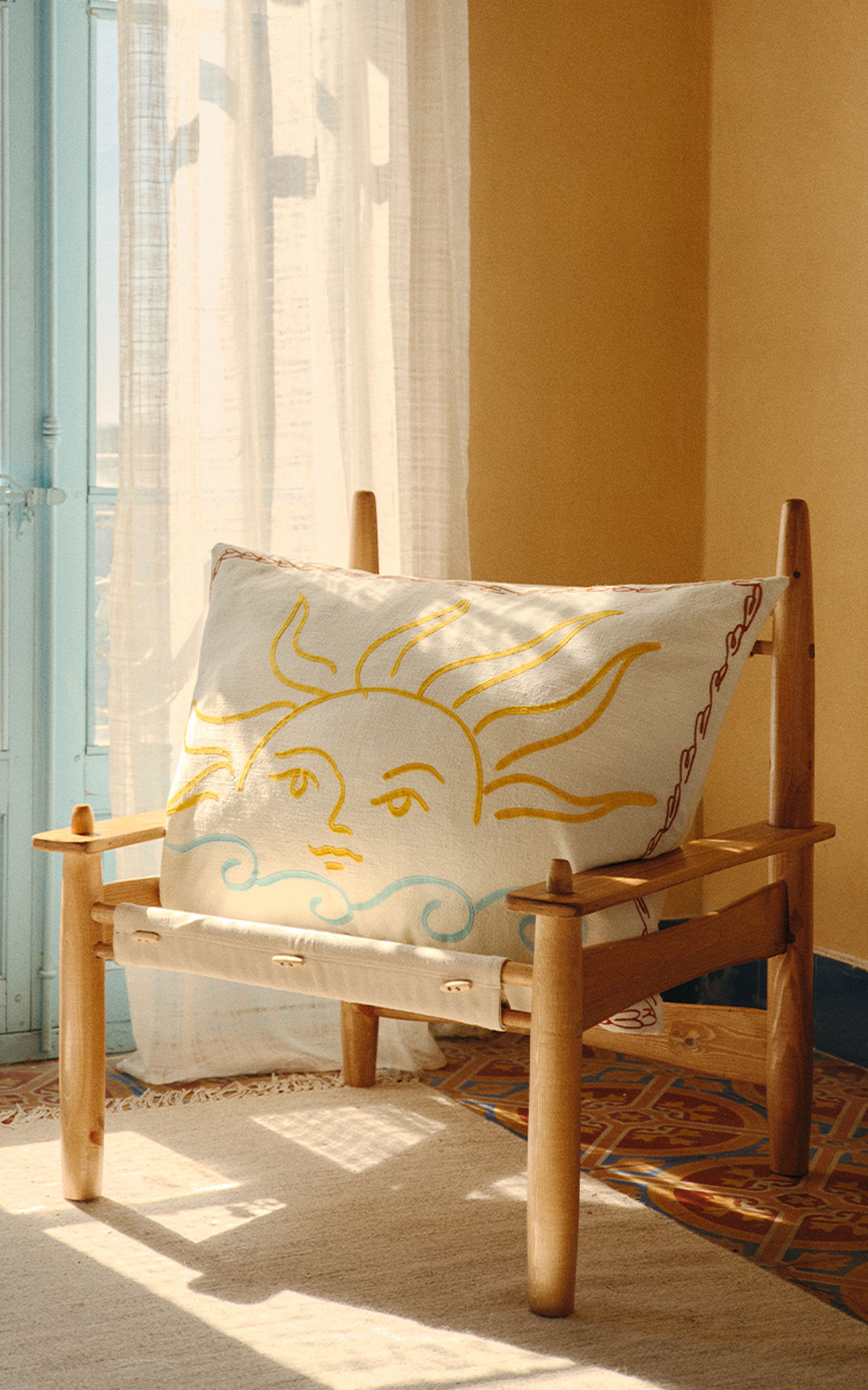 Une chaise en bois avec un coussin sur le thème du soleil, dans une pièce baignée de lumière, avec des rideaux clairs et un sol à motifs.