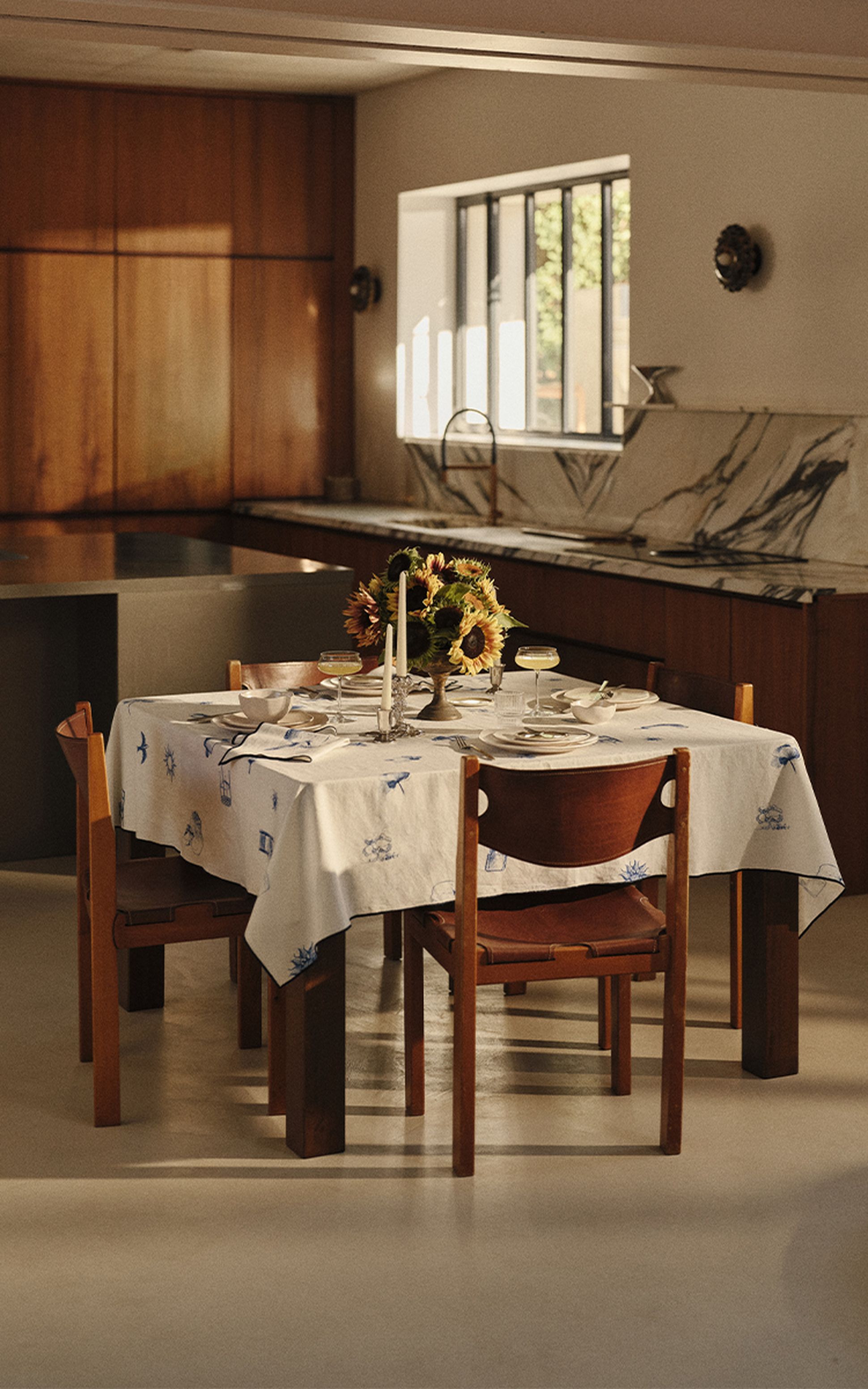 Une table à manger dressée pour quatre personnes, avec une nappe blanche, des tournesols, des bougeoirs et de la vaisselle, dans une cuisine moderne baignée d'une lumière chaleureuse.
