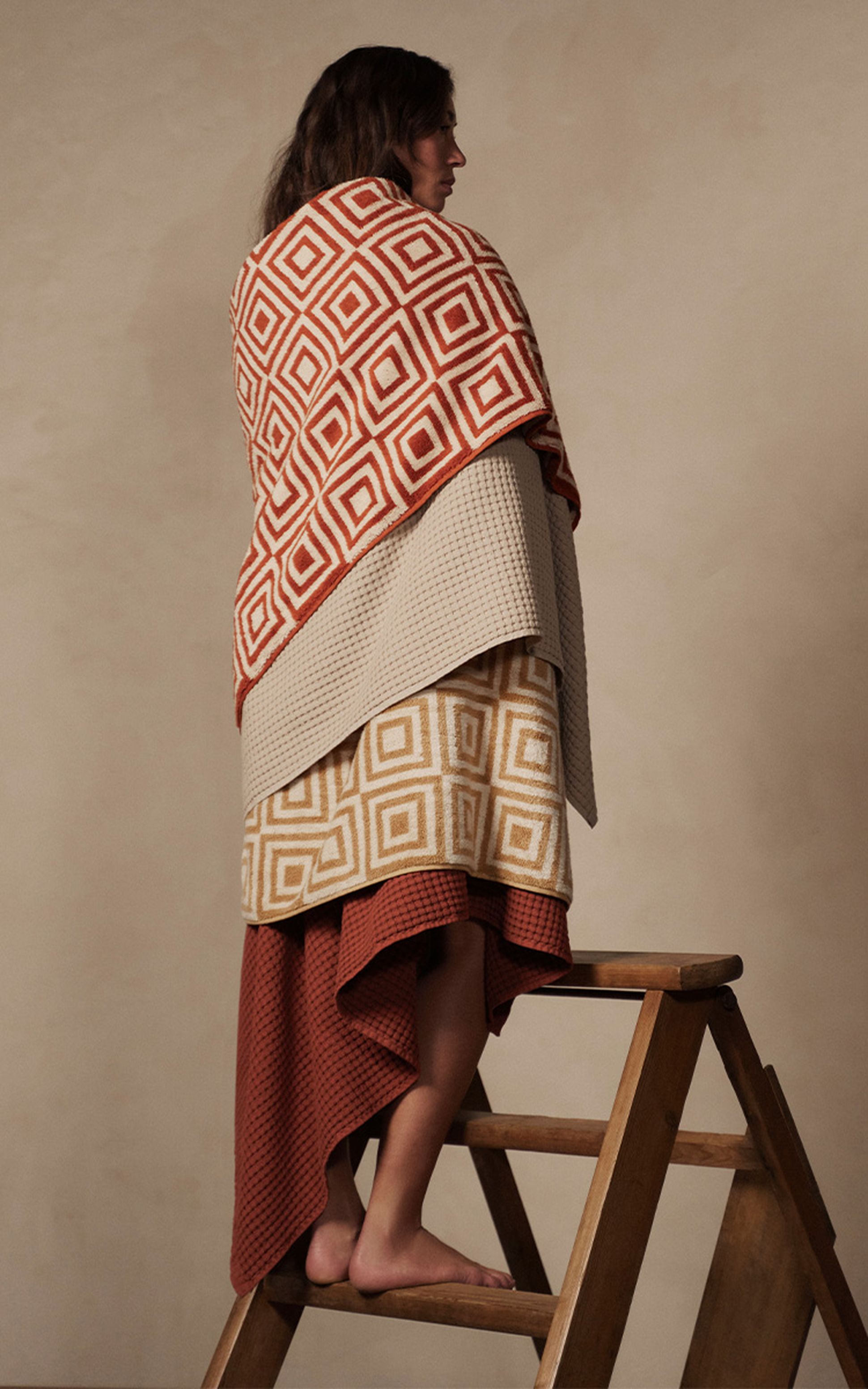 Une personne enveloppée dans des couvertures à motifs géométriques se tient debout sur une échelle en bois devant un fond neutre.