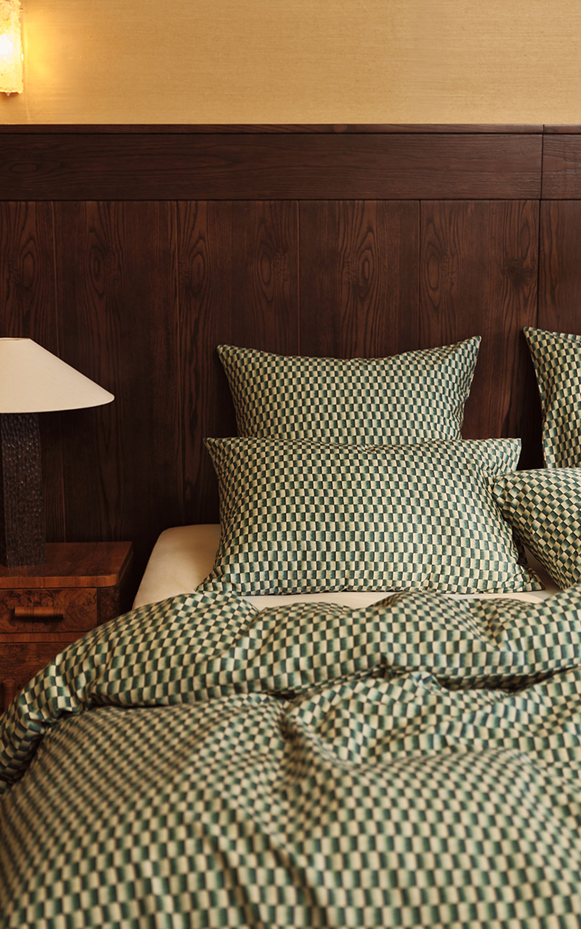 Une chambre douillette avec un lit double, une parure de lit à carreaux, une tête de lit en bois, une lampe de chevet et une fenêtre aux rideaux laissant passer une douce lumière.