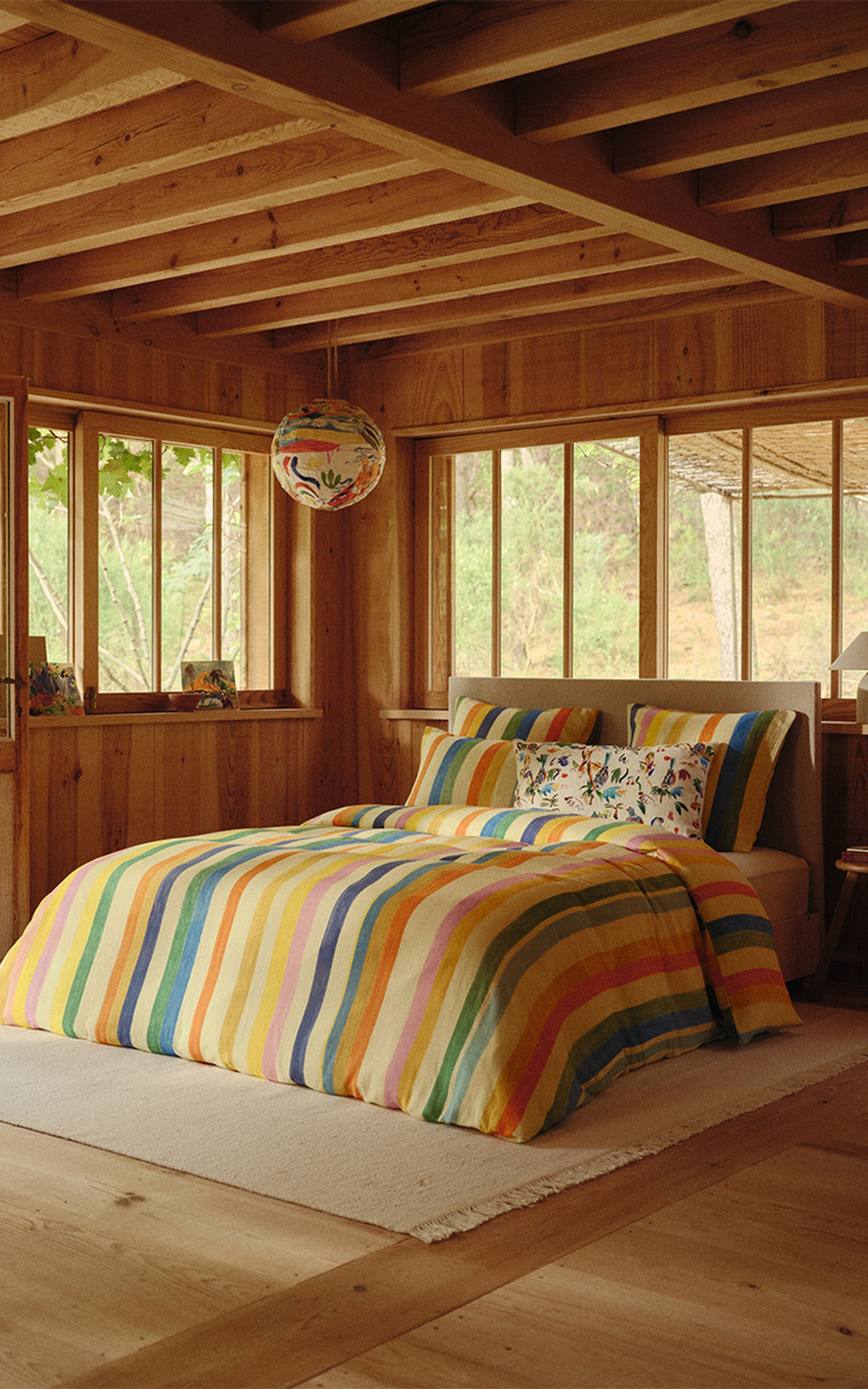Une chambre chaleureuse en bois dotée de grandes fenêtres, avec un lit orné d'une parure de lit à rayures colorées et d'oreillers décoratifs, sous une lanterne suspendue aux couleurs vives.