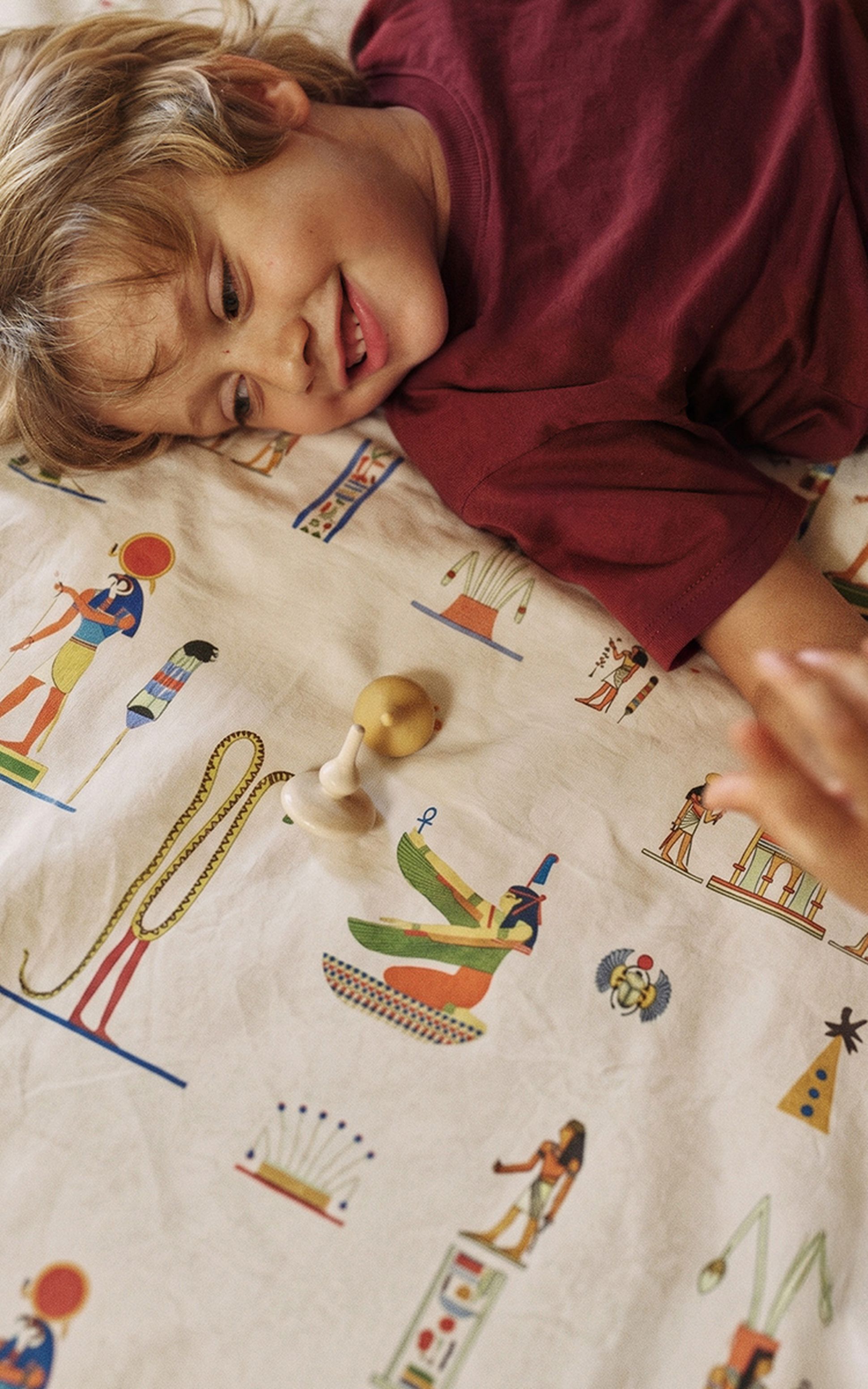 Un enfant allongé sur un drap aux motifs égyptiens, souriant et tendant la main vers une toupie en bois.