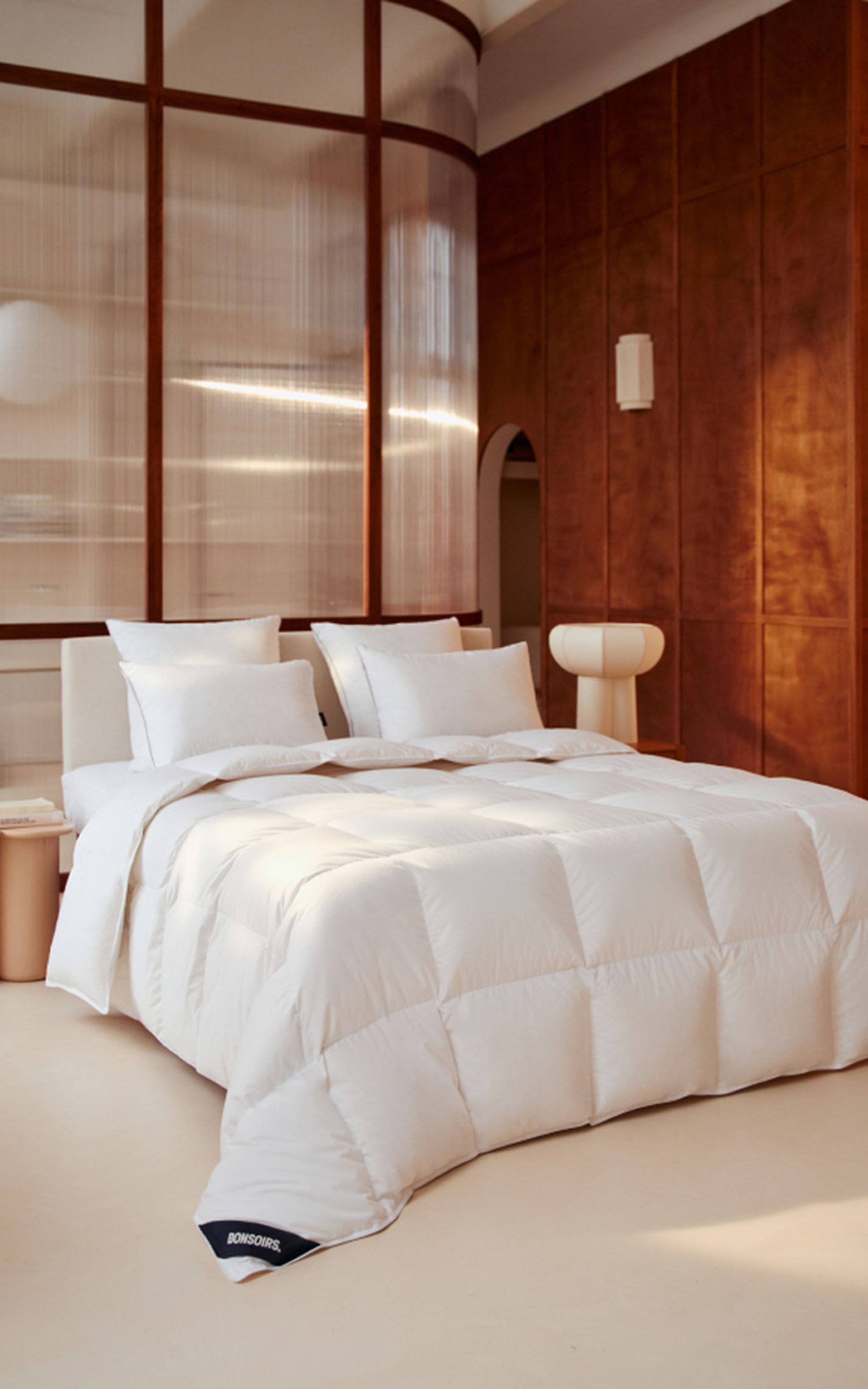 A modern bedroom with a white bed, three pillows, and a wood-accented wall. Natural light filters through a translucent partition.