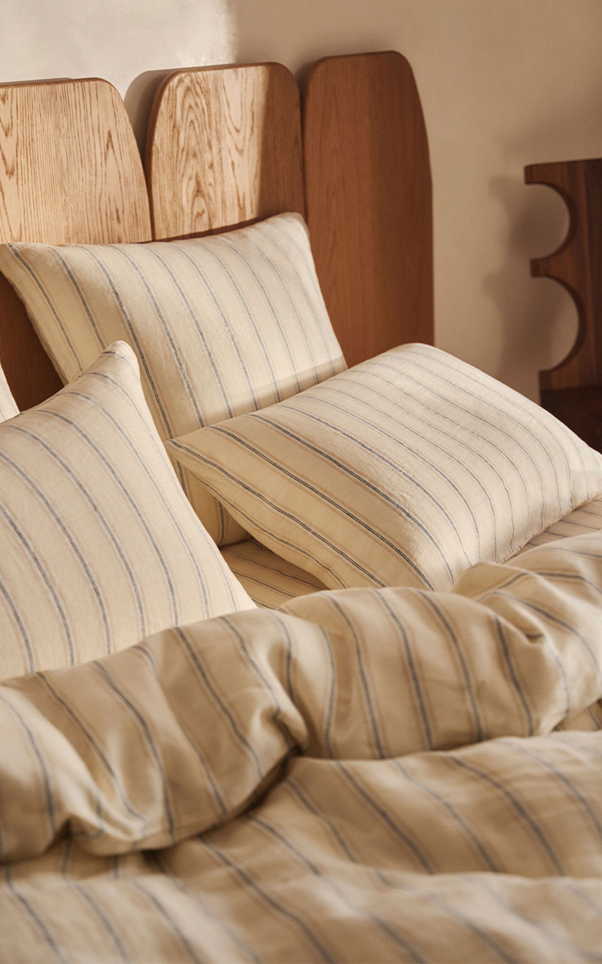 Une parure de lit beige rehaussée de fines rayures bleues, posée sur un lit à tête de lit en bois, baignée d'une douce lumière naturelle.