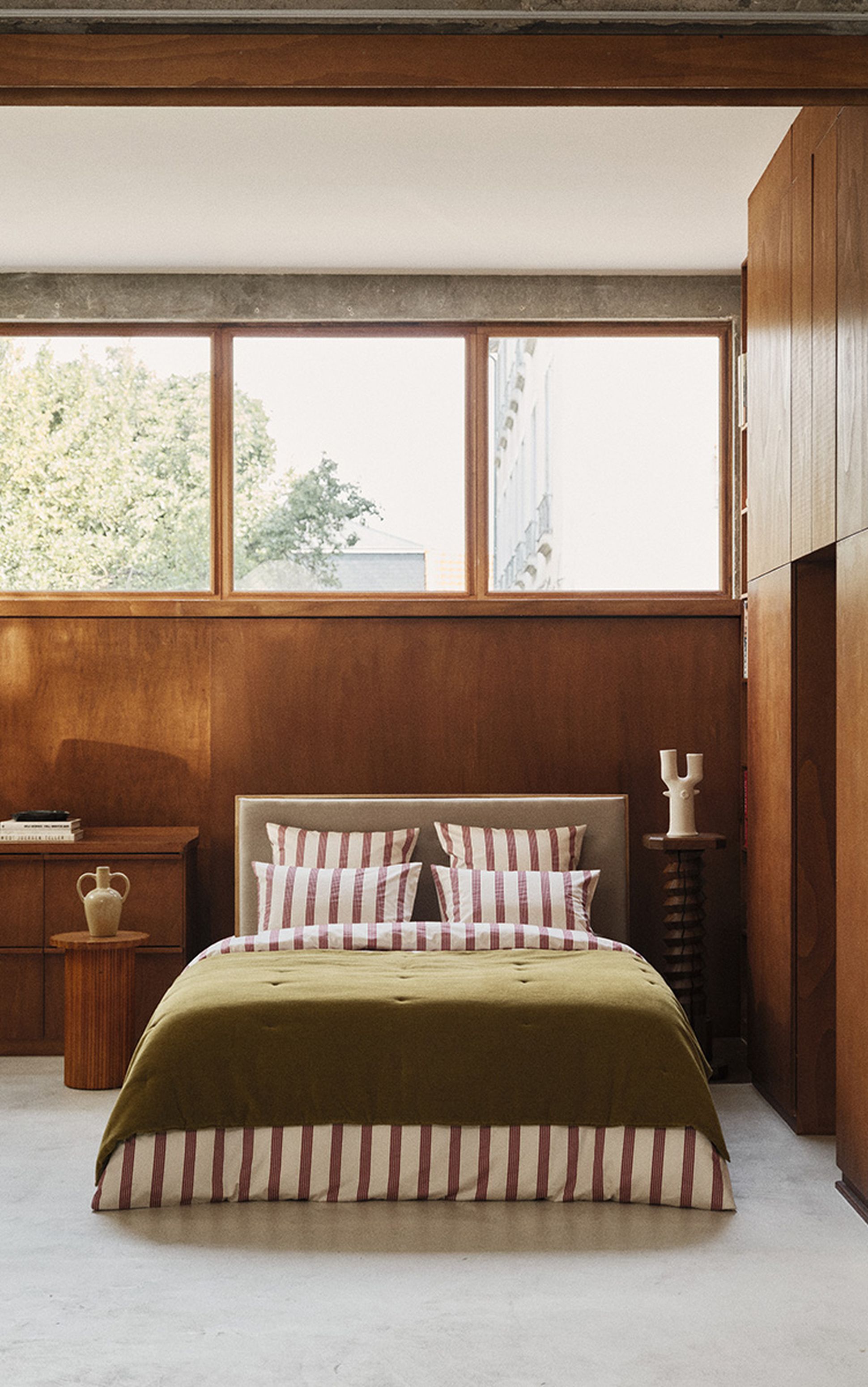 Une chambre douillette avec un lit orné d'oreillers rayés et d'une couverture verte, des tables de chevet en bois, des vases, des livres et un éclairage tamisé, le tout devant un mur en bois.
