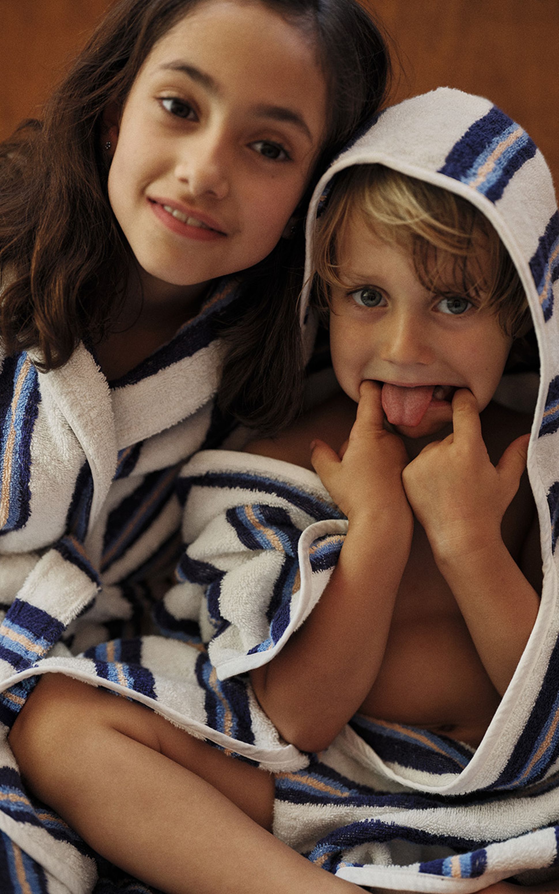 Deux enfants en serviette rayée, l'un souriant et l'autre tirant la langue d'un air espiègle.