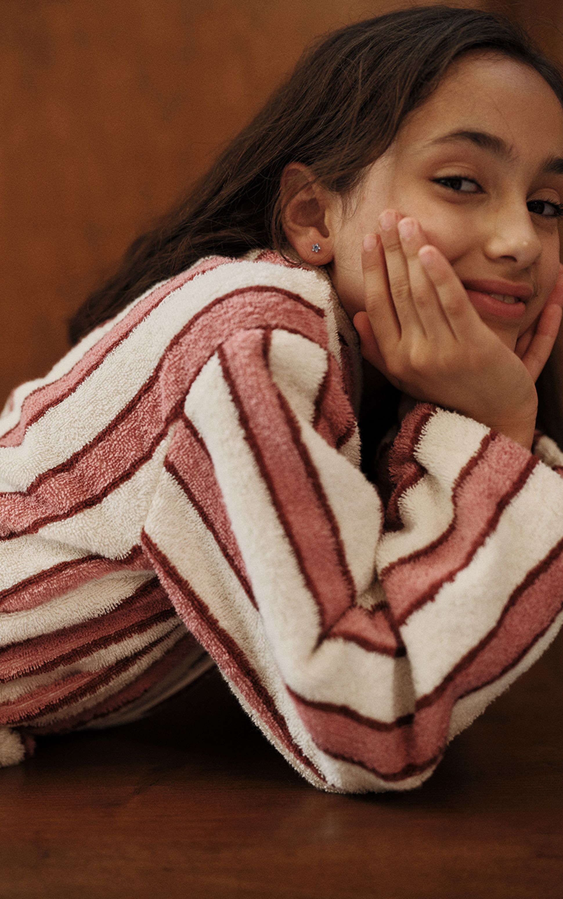 Une jeune fille souriante, les mains posées sur les joues, vêtue d'un pull rayé rose et blanc, appuyée contre une table en bois.
