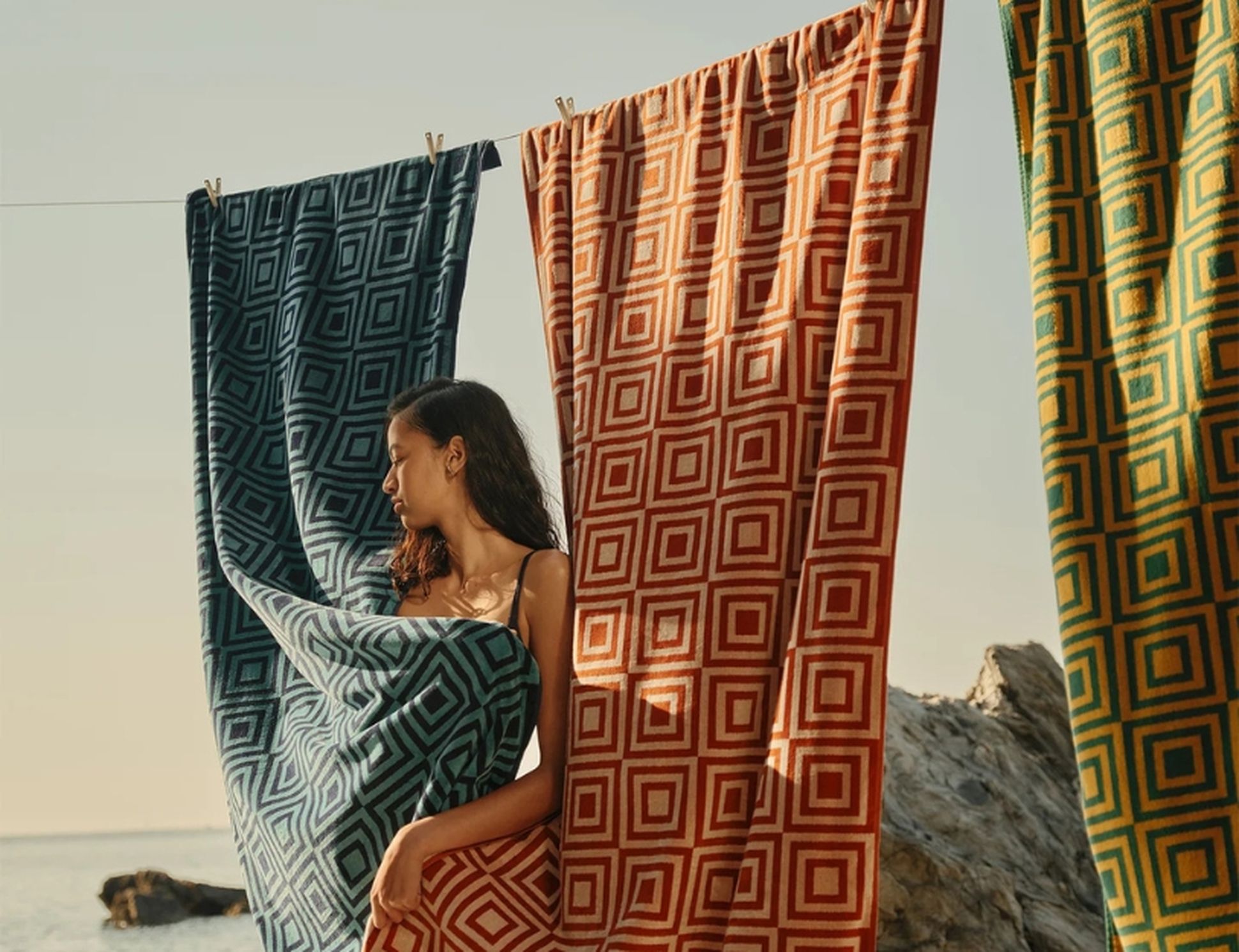 Une femme en bikini se tient au bord de la mer, encadrée par trois grandes serviettes à motifs bleues, rouges et jaunes.