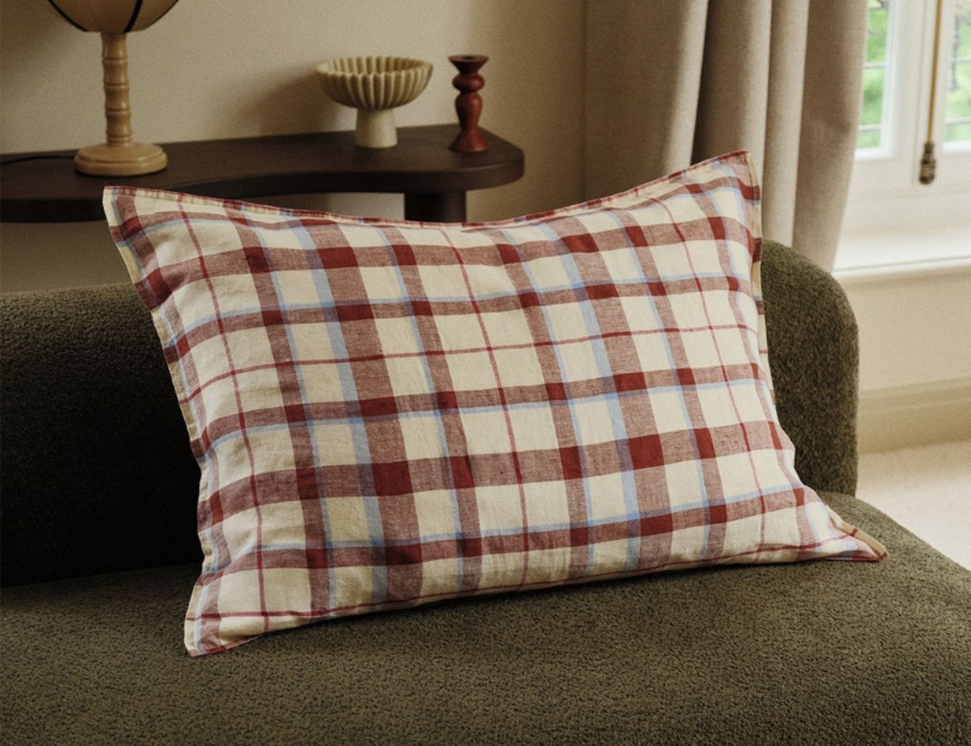 Un coussin à carreaux, avec des rayures rouges, bleues et beiges, posé sur un canapé vert foncé dans une pièce chaleureuse agrémentée d'éléments de décoration en bois.