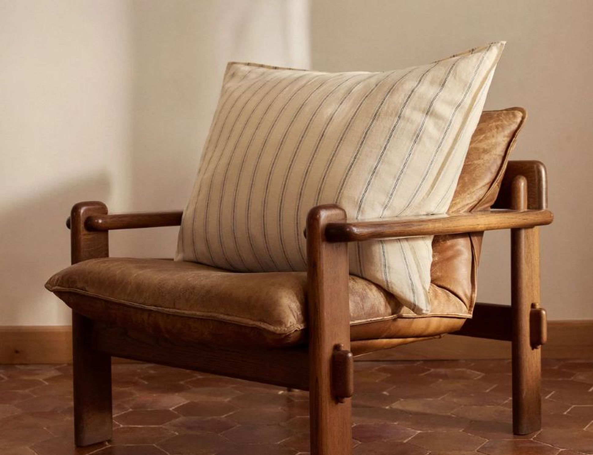 Chaise longue en bois avec un coussin en cuir marron et un oreiller blanc rayé, placée devant un mur clair et un sol carrelé.
