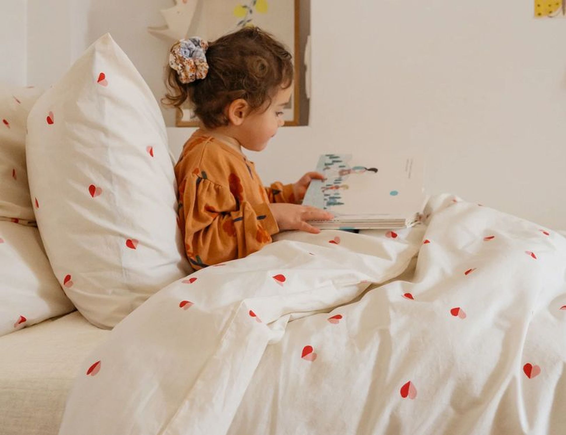 Une enfant vêtue d'une robe marron est assise sur un lit aux draps à motifs de cœurs et lit un livre.